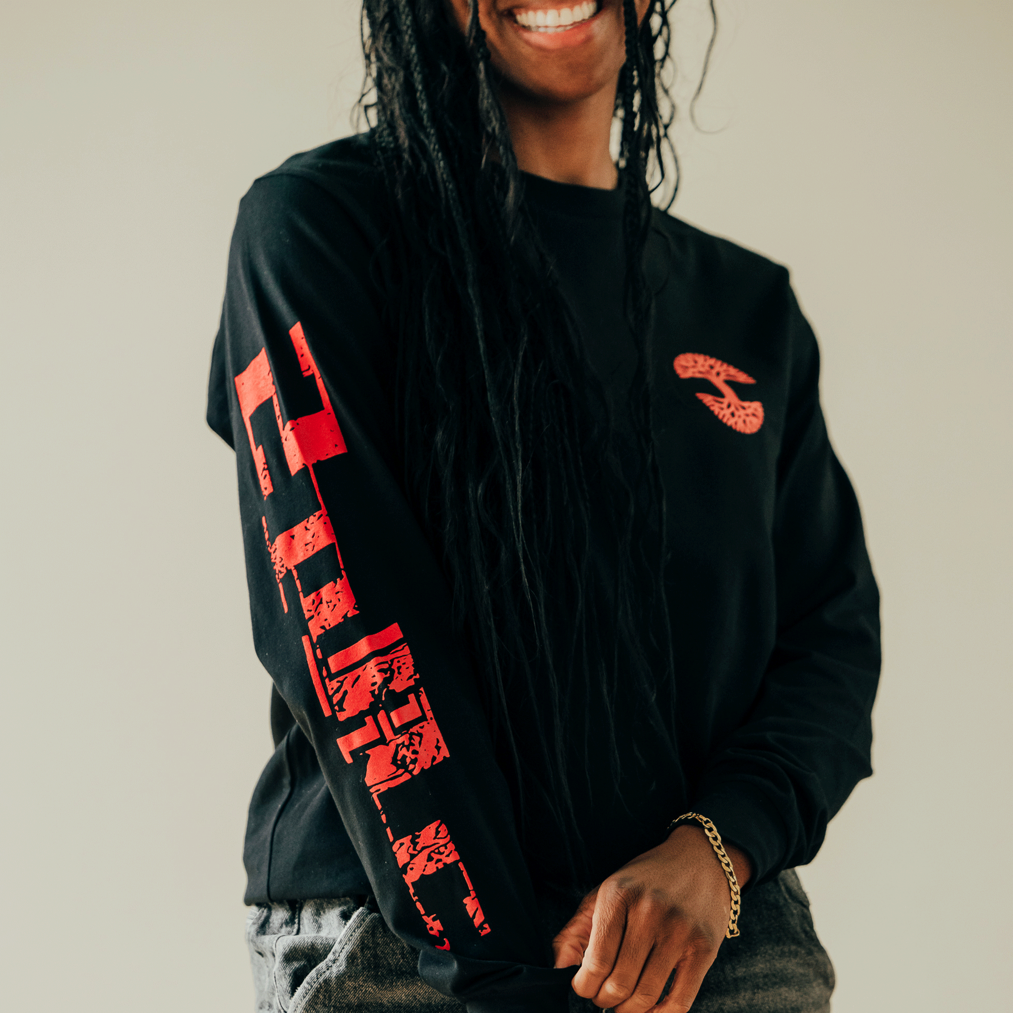 The person, sporting long braided hair, is wearing the "You Got To KYHU LS Tee" by Oaklandish. This black sweatshirt showcases a red snake emblem on the chest and bold red lettering down the sleeve. Reflecting the Oaklandish x Tupac collaboration, they have a slight smile while casually holding the bottom of the sweatshirt against a neutral background.