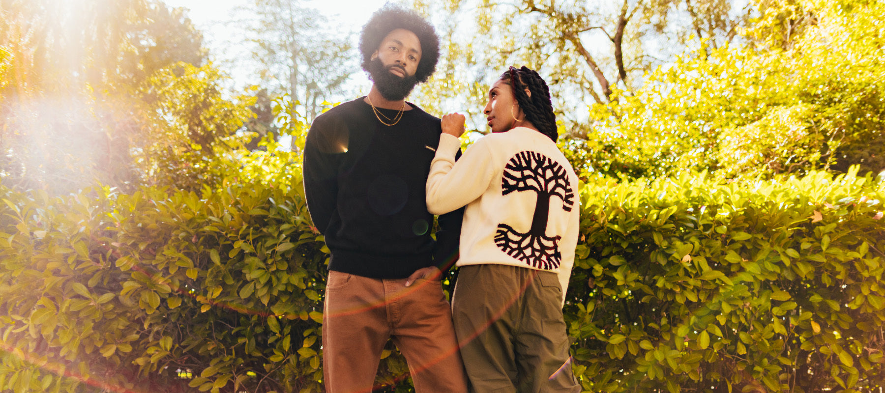 Male and female model wearing Oaklandish logo knit sweaters outside in front of shrubs.