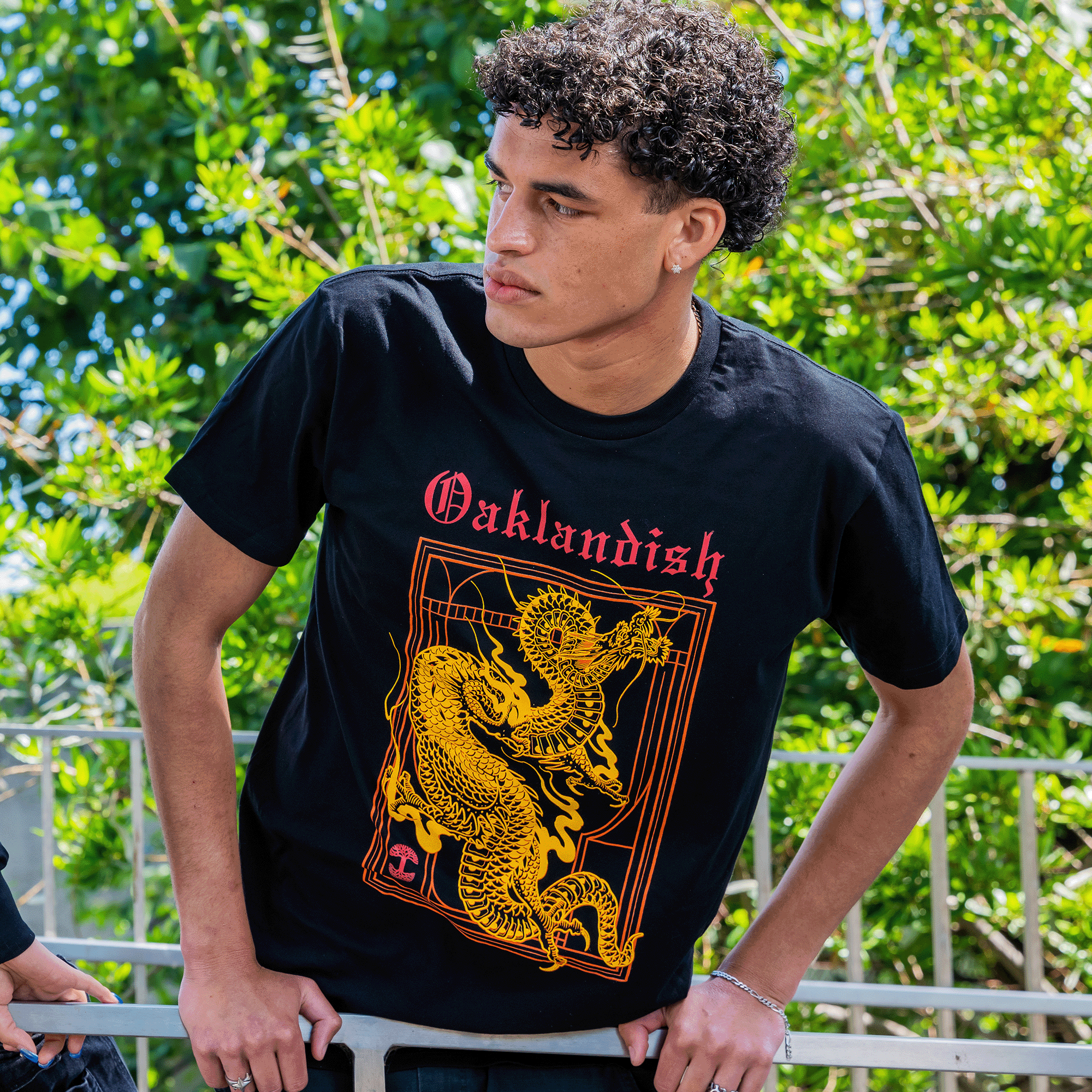 A person with curly hair leans against a railing outdoors, looking to the side. They wear the Oaklandish Prosperity Tee, featuring a classic fit with a red "Oaklandish" logo and an intricate yellow dragon design symbolizing prosperity. The lush green foliage backdrop complements their look, accessorized stylishly with a bracelet.