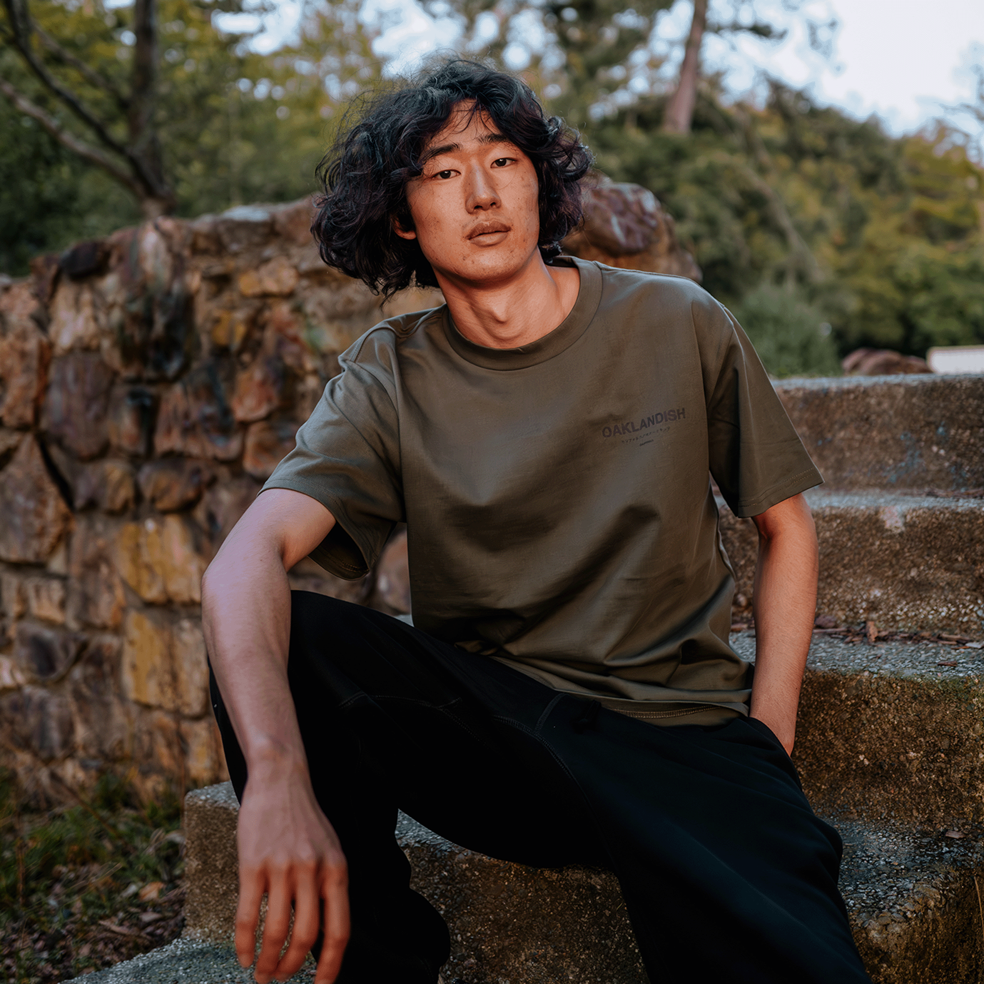 A person with curly hair sits on outdoor stone steps, wearing the Oaklandish Modern Type Heavy Tee in green along with black pants. In the background are a stone wall and lush greenery, with warm late afternoon lighting enhancing a relaxed and casual atmosphere.