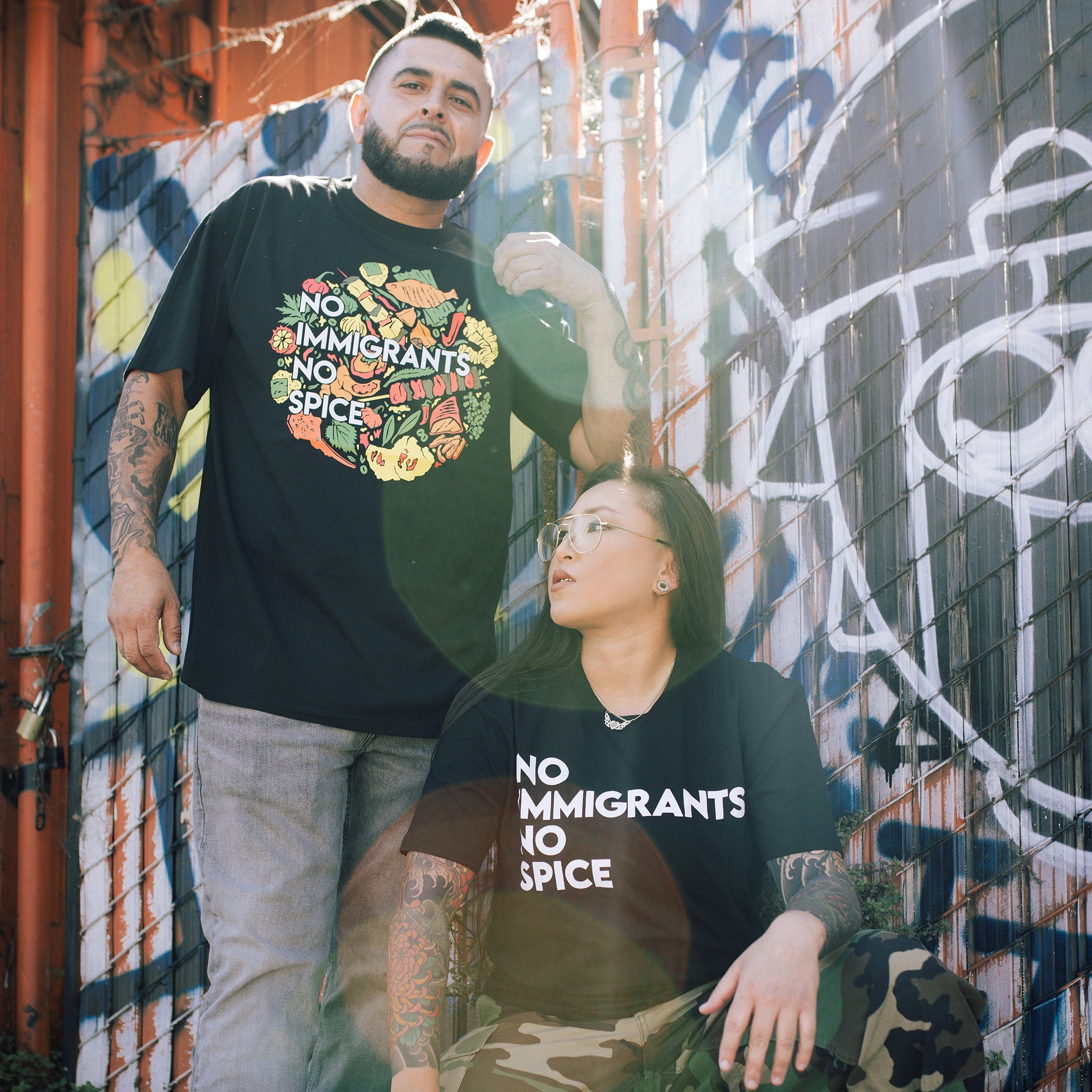 Two people pose against a graffiti wall. The man, standing, wears an Oaklandish NINS Classic Tee with "No Immigrants No Spice" text. The woman, seated in solidarity, wears glasses and a matching tee. Both have tattoos as sunlight creates lens flares in the image.