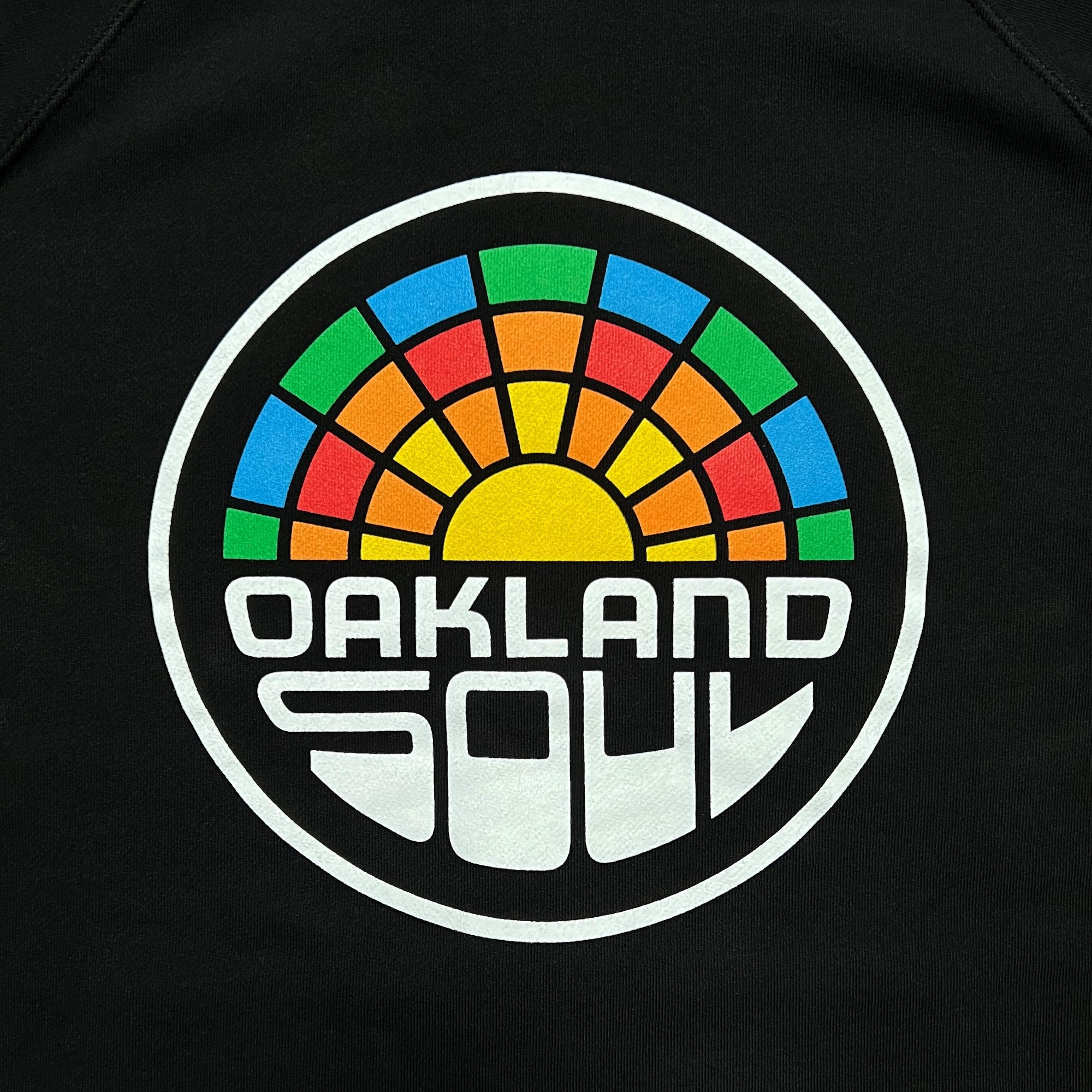 The Oakland Soul Logo Zip, from Oakland Roots SC, features a circular logo with an outer black ring containing the words "Oakland Soul SC" in stylized white text. Inside the circle, a bright yellow half-sun is depicted with colorful rainbow-like rays in blue, green, and red. This vibrant retro design against a dark background creates the perfect emblem for your unisex zip hoodie supporting social good.