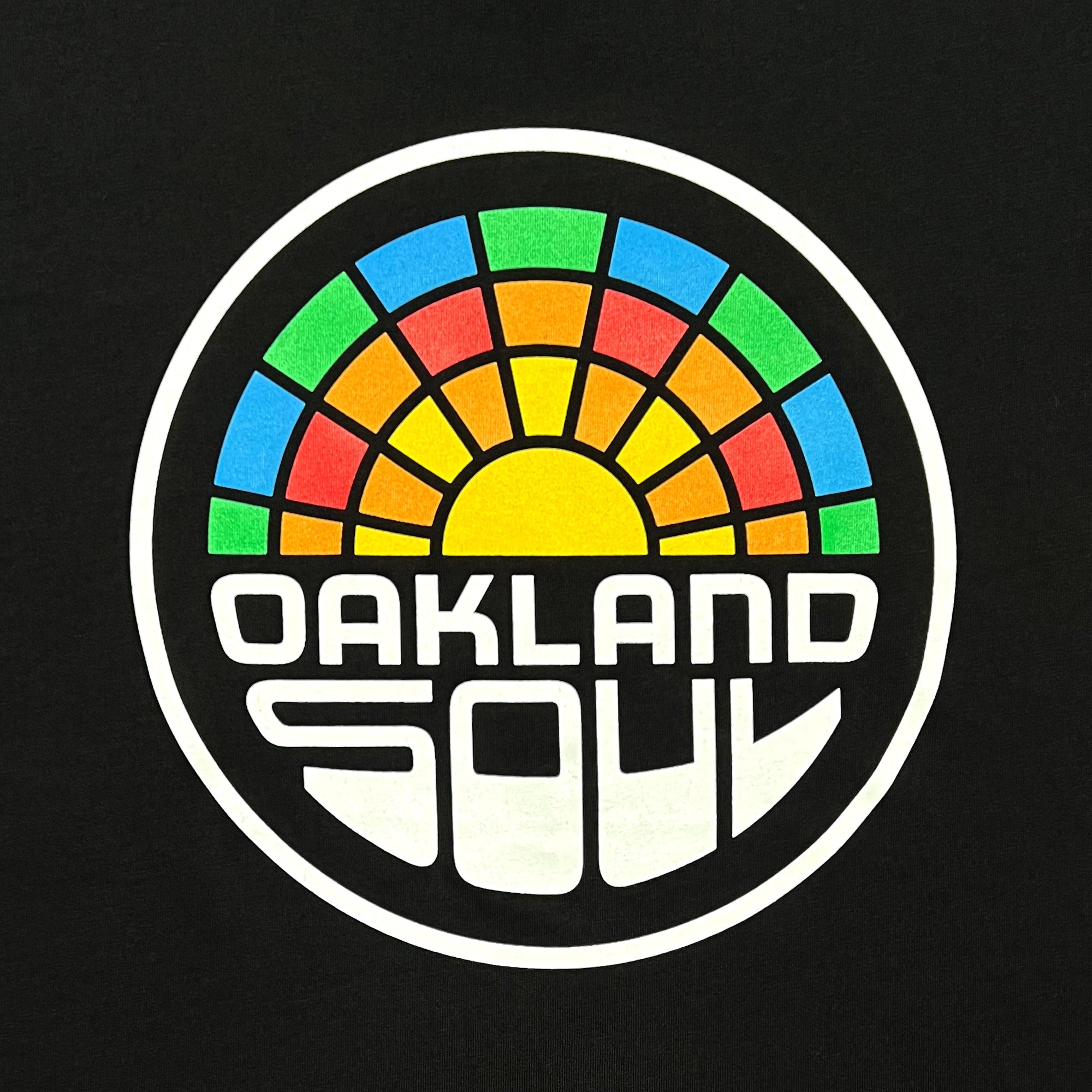 The Oakland Soul Logo Tee, by Oakland Soul, features a black background with a circular logo that includes a multicolored, stylized sunrise representing the Oakland Soul women's soccer team in the USL W-League. The top half of the design is segmented into green, orange, yellow, and blue sections. Below the sunrise, bold white text reads "OAKLAND SOUL." The overall aesthetic is reminiscent of a stained glass window.