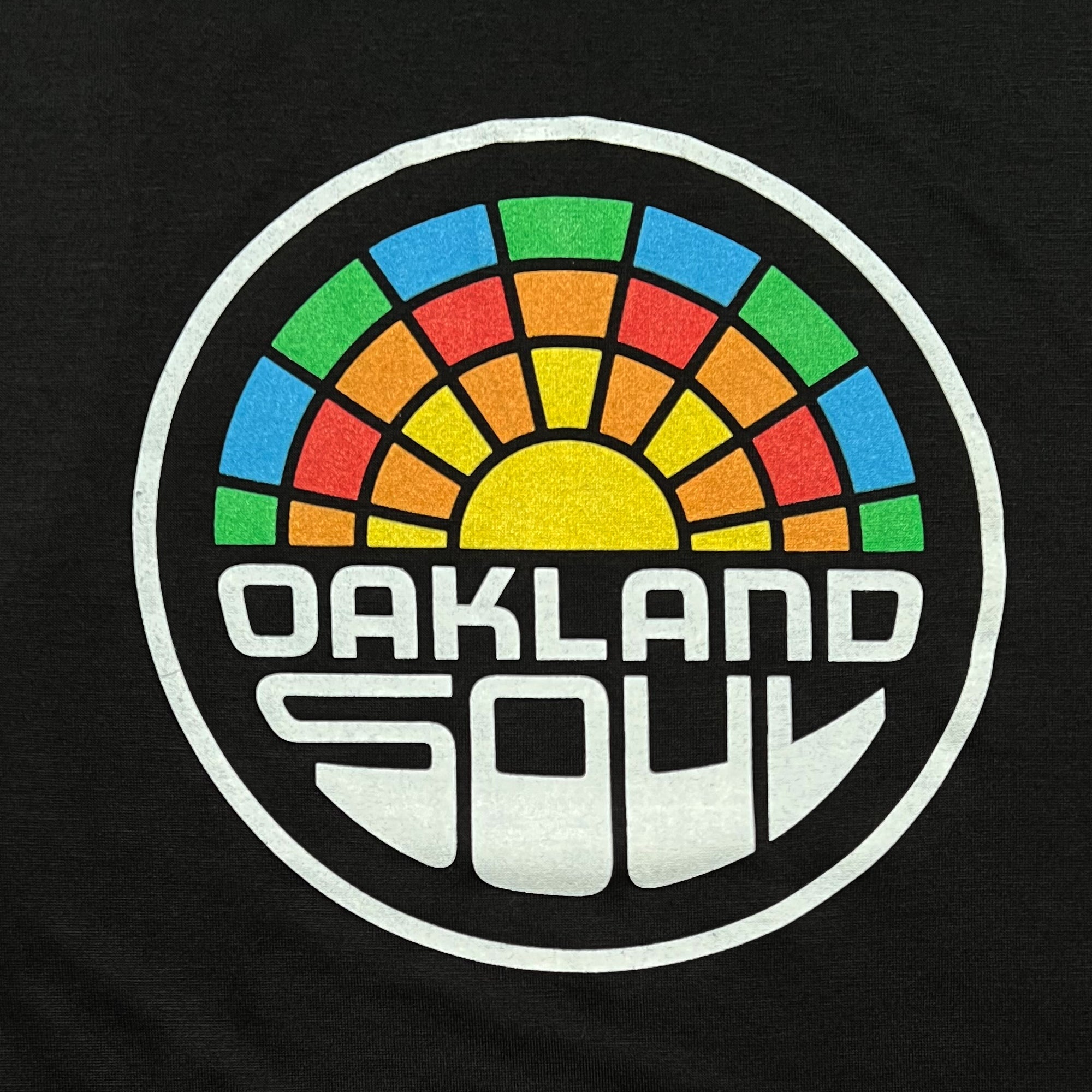 A circular logo against a dark background features a stylized image of a radiant sun with multicolored rays in green, blue, red, and yellow. Below the sun are the words "OAKLAND SOUL" in bold white letters, encapsulating the vibrant spirit of women's soccer in the USL W-League. The design is enclosed within a thick white circle on the Women's Oakland Soul Logo Tank by Oakland Soul.