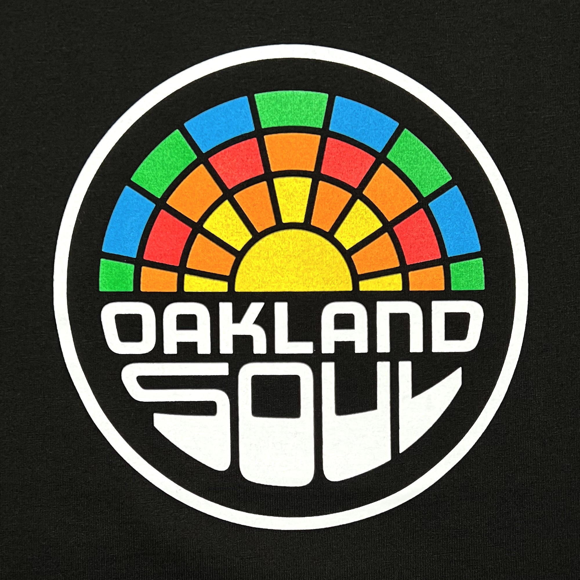 The Women's Oakland Soul Logo Tee by Oakland Roots SC features a circular logo with bold, white, stylized text reading "OAKLAND SOUL SC." Above the text is a semicircular mosaic pattern in multiple colors, evoking the imagery of a sunrise or sunset. The logo stands out against the black background of this classic fit t-shirt, available in women's sizing.