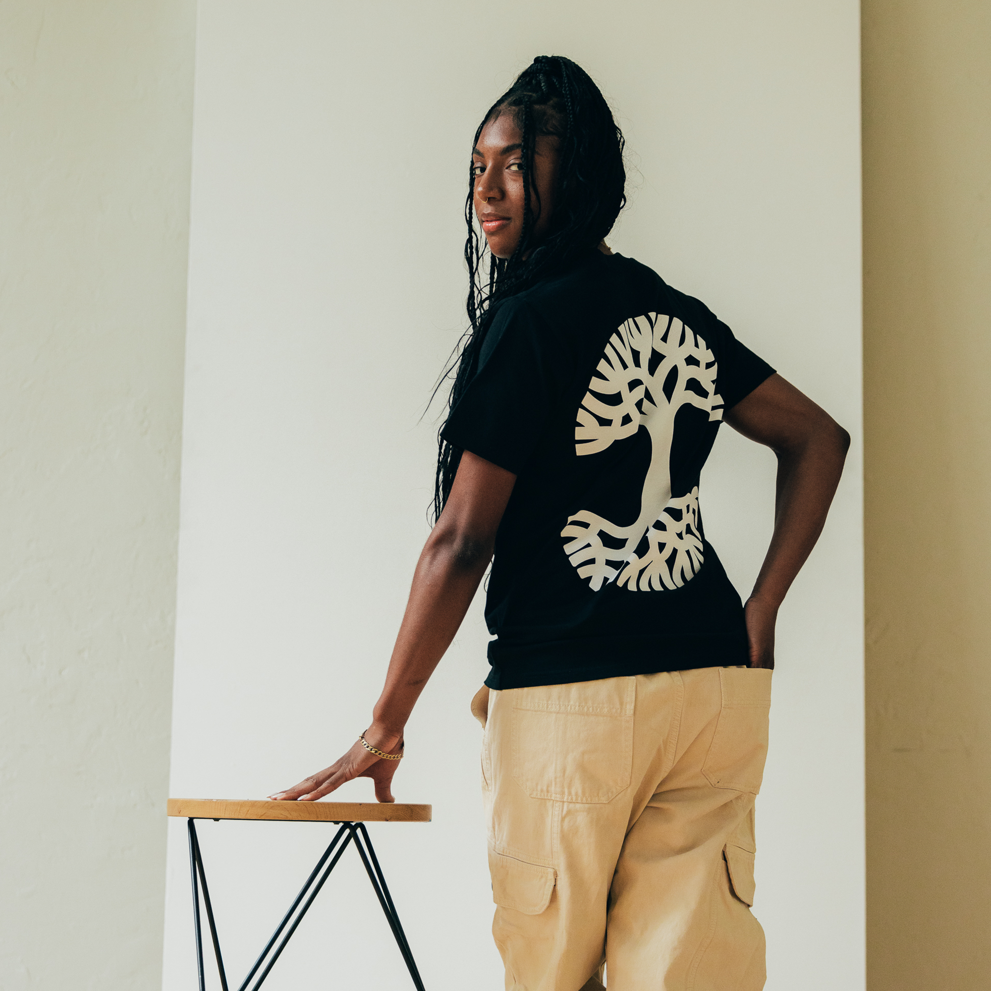 A person with long braided hair stands sideways wearing the PacLogo Tee, a black T-shirt by Oaklandish featuring a tree-like design on the back. They lean on a small folding table with their left hand, while their beige pants and the plain light-colored wall complete this hip-hop inspired look.