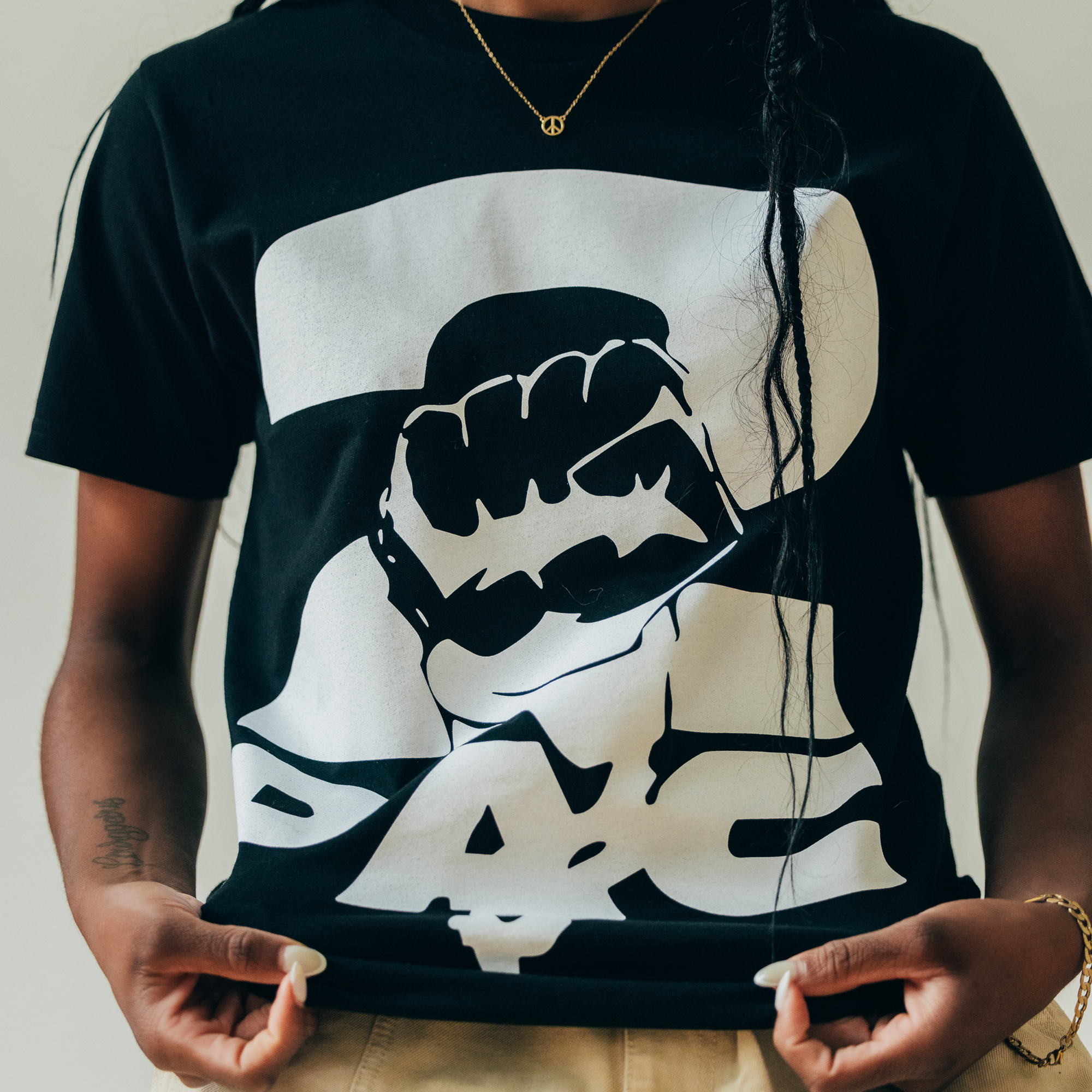 Wearing the PacLogo Tee from Oaklandish, featuring a black t-shirt with a white graphic design of a raised fist in the center and "PAC" boldly spelled at the bottom, this person exudes hip-hop vibes. Sporting long braided hair and accessorized with a necklace with a small round pendant, they embody an Oaklandish style while holding the shirt at its sides.