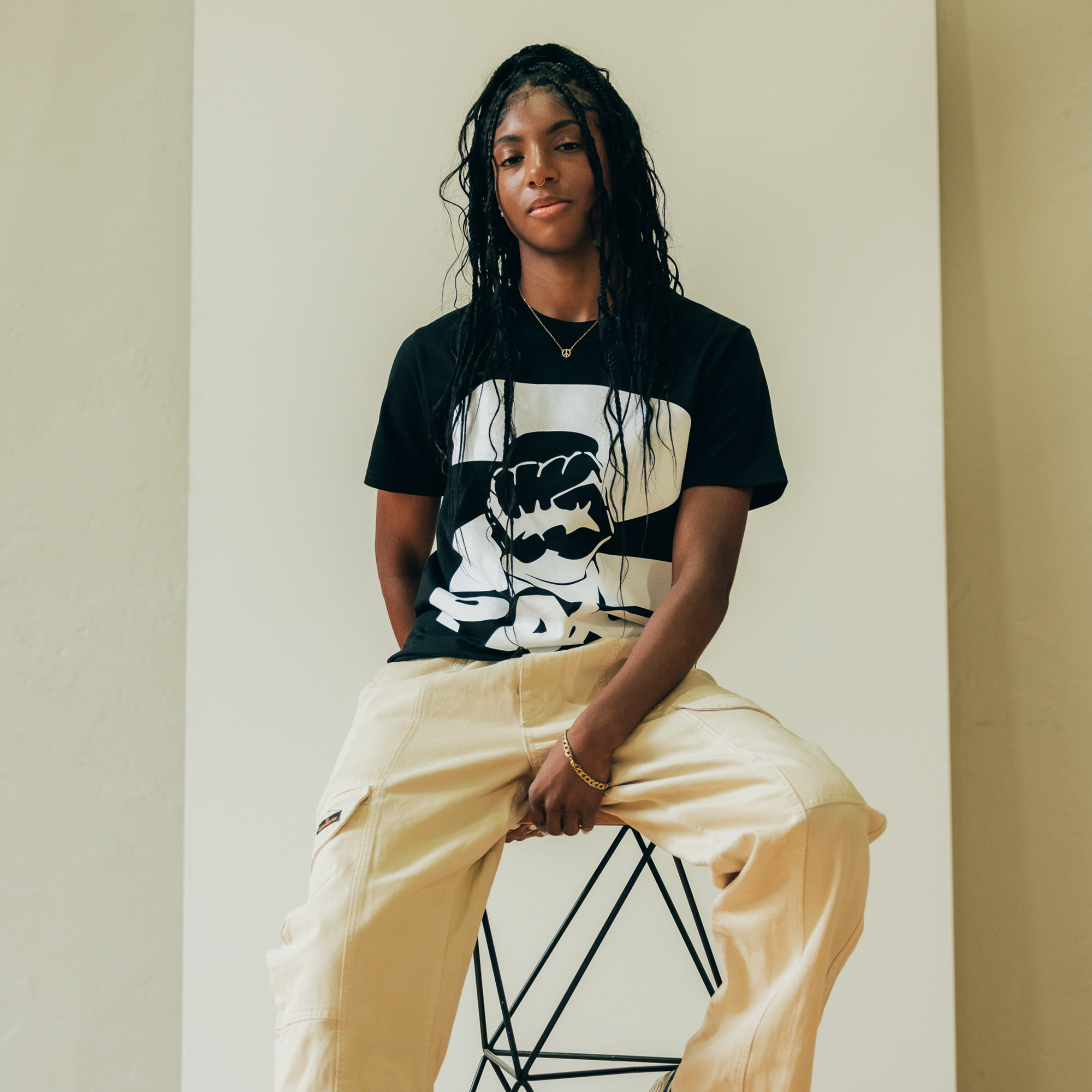 A person with long, wavy hair wearing an Oaklandish PacLogo Tee and light beige pants sits casually on a modern geometric stool. The neutral ivory background complements their confident demeanor, exuding a relaxed, stylish vibe that channels hip-hop cool.