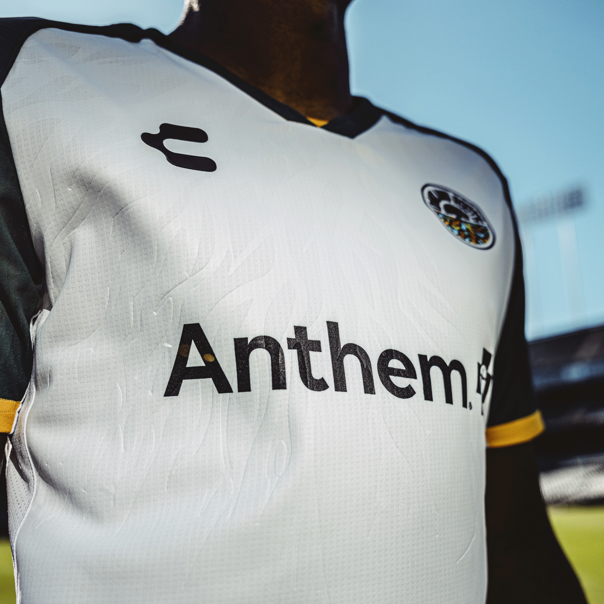 Close-up of someone wearing the "Unisex Oakland Roots SC 2025 Second Kit ft. Anthem Blue Cross with JÜRGEN 25," showcasing "Anthem" and an Embossed Roots Design on a white and dark green jersey, with a dark collar and trim, against a blurred stadium backdrop under clear blue skies.