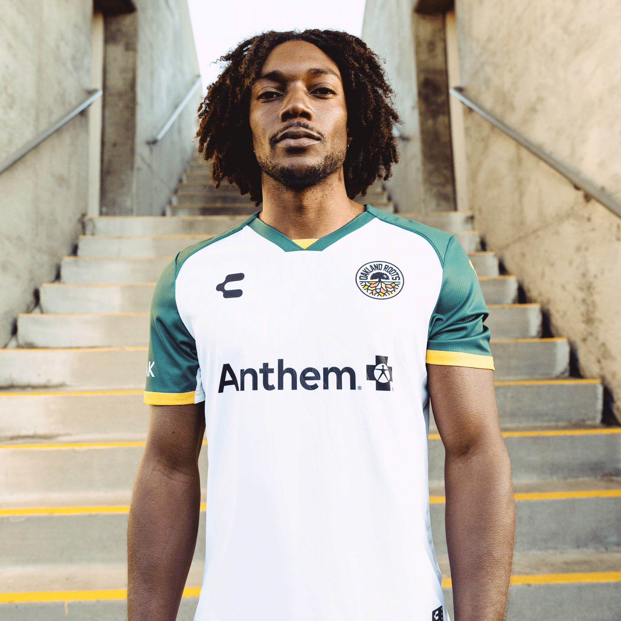 A man with shoulder-length curly hair stands on a concrete staircase, wearing the Custom Unisex Oakland Roots SC 2025 Second Kit featuring Anthem Blue Cross. The jersey reflects Oakland sports culture, adorned with an embossed Roots design and round emblem on the right side. Confidently, he gazes at the camera.