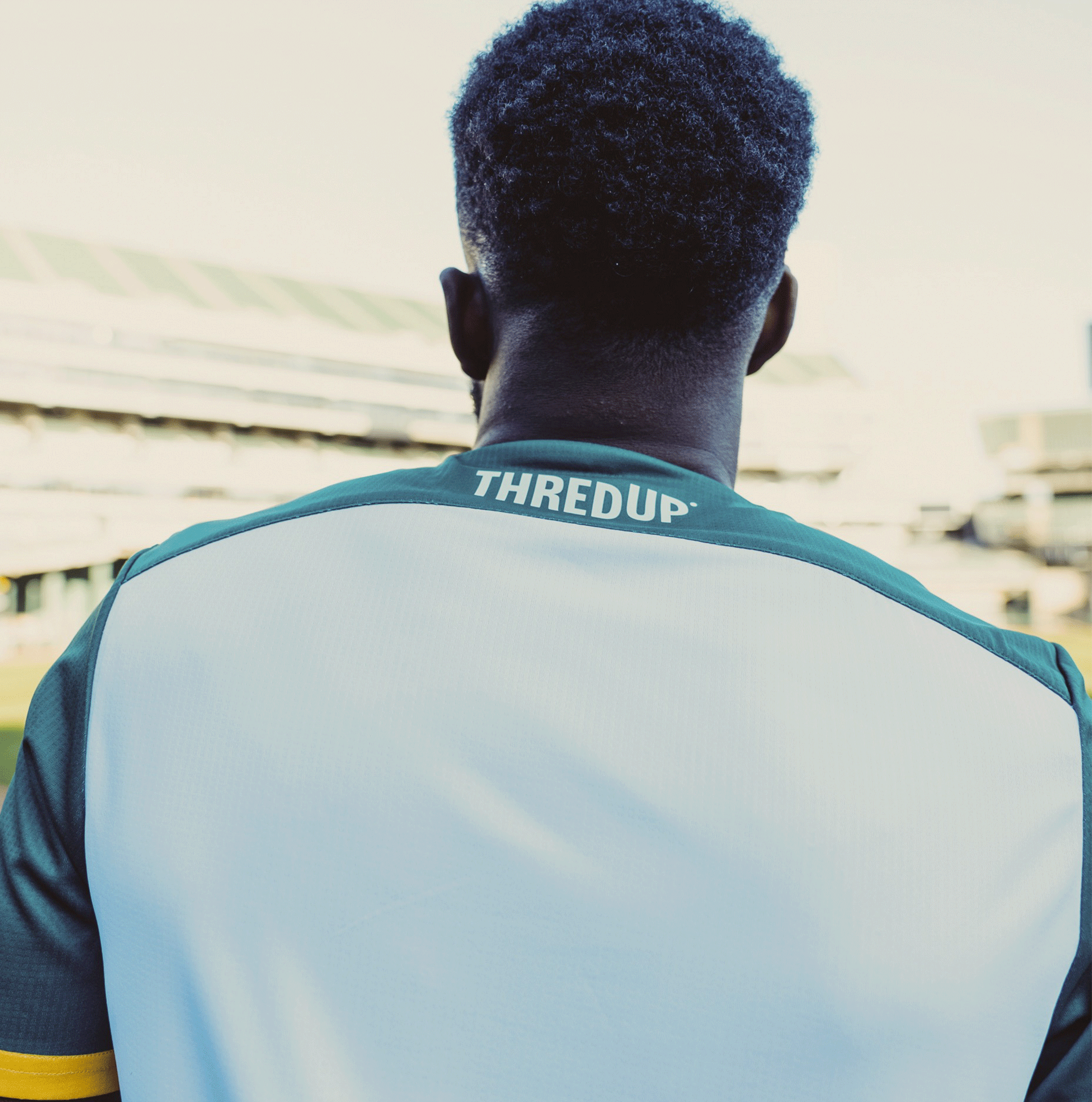 In an empty stadium, someone wears the Unisex Oakland Roots SC 2025 Second Kit with "THREDUP" on the back, looking out where Oakland Roots SC made history. The clear sky above Coliseum Coordinates adds a serene anticipation.