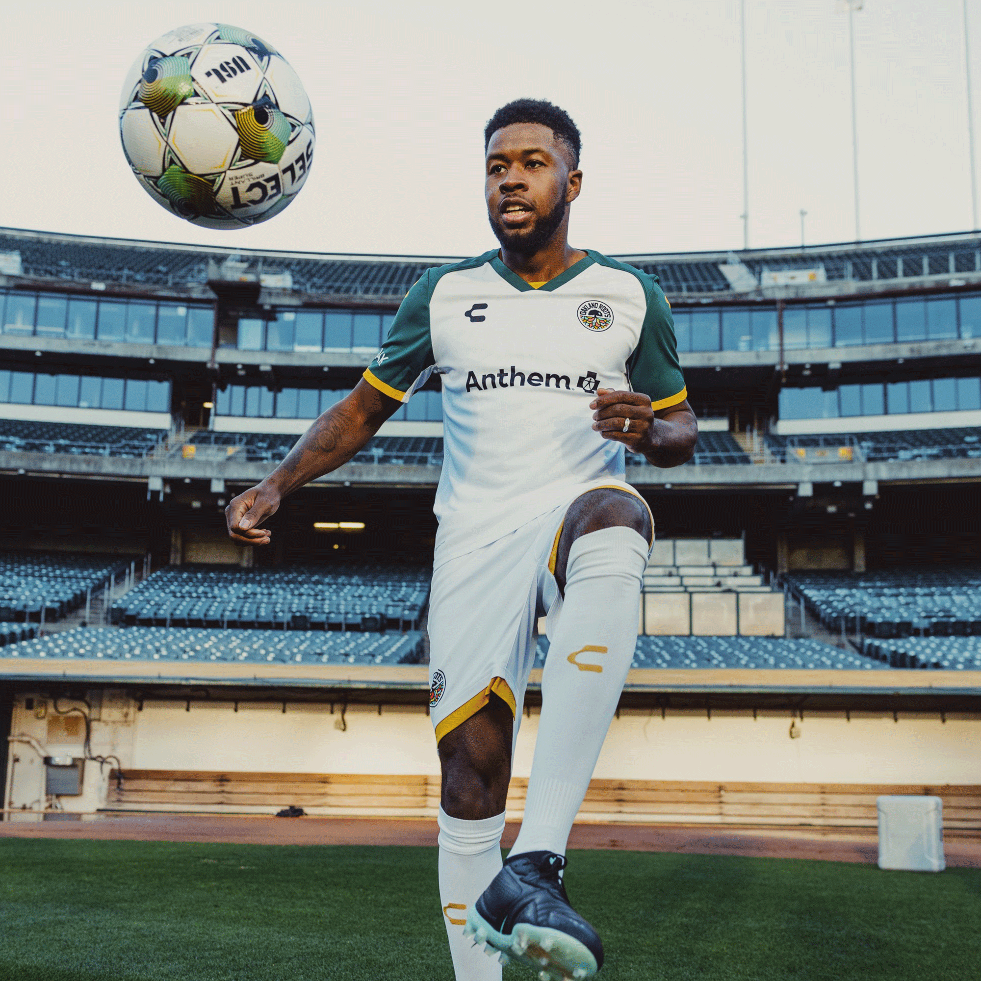 A soccer player in a white and green "Unisex Oakland Roots SC 2025 Second Kit ft. Anthem Blue Cross" uniform juggles a ball on his knee on the grassy field of an empty stadium, spotlighted amid rows of seats, capturing the essence of Oakland sports history.