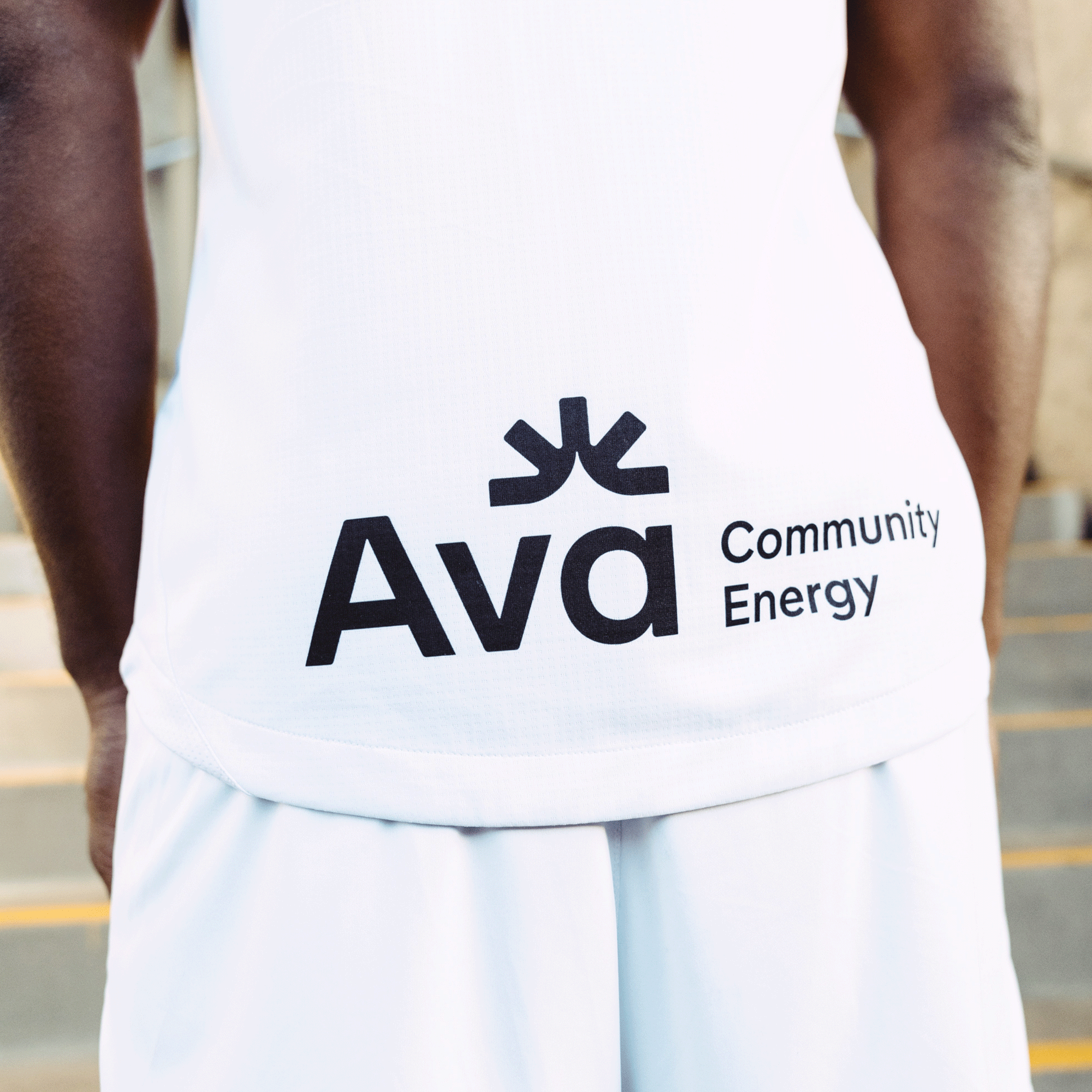 Close-up of a person wearing the Custom Unisex Oakland Roots SC 2025 Second Kit ft. Anthem Blue Cross with "Ava Community Energy" printed in black alongside an abstract logo. Blurred stone steps form the backdrop, highlighting just the lower torso and capturing community essence.