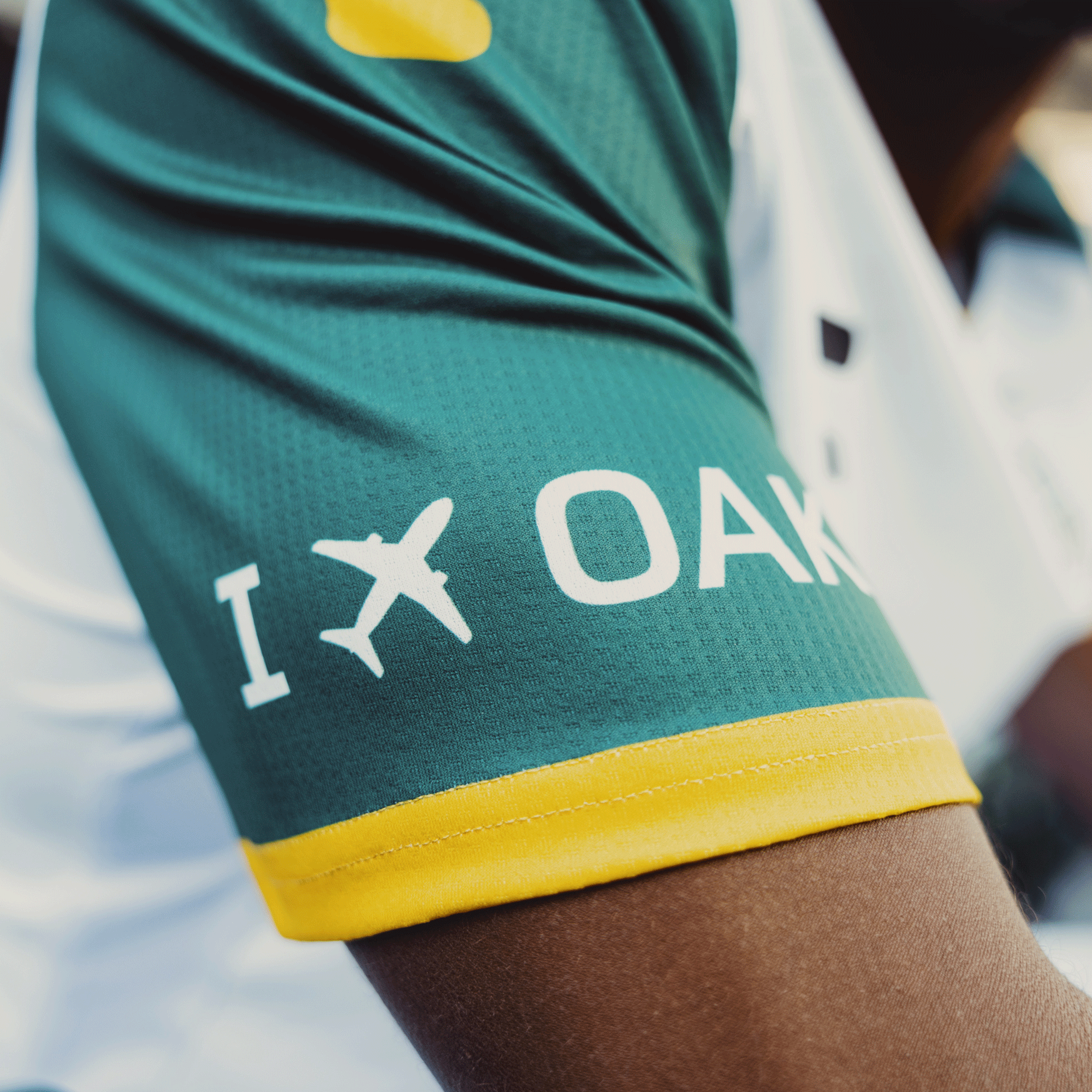 Close-up of someone wearing the Unisex Oakland Roots SC 2025 Second Kit featuring Anthem Blue Cross, displaying "I [airplane icon] OAK" in white with yellow trim on a green jersey sleeve, reflecting sports history and Oakland Roots. A blurred background completes this sporty image.