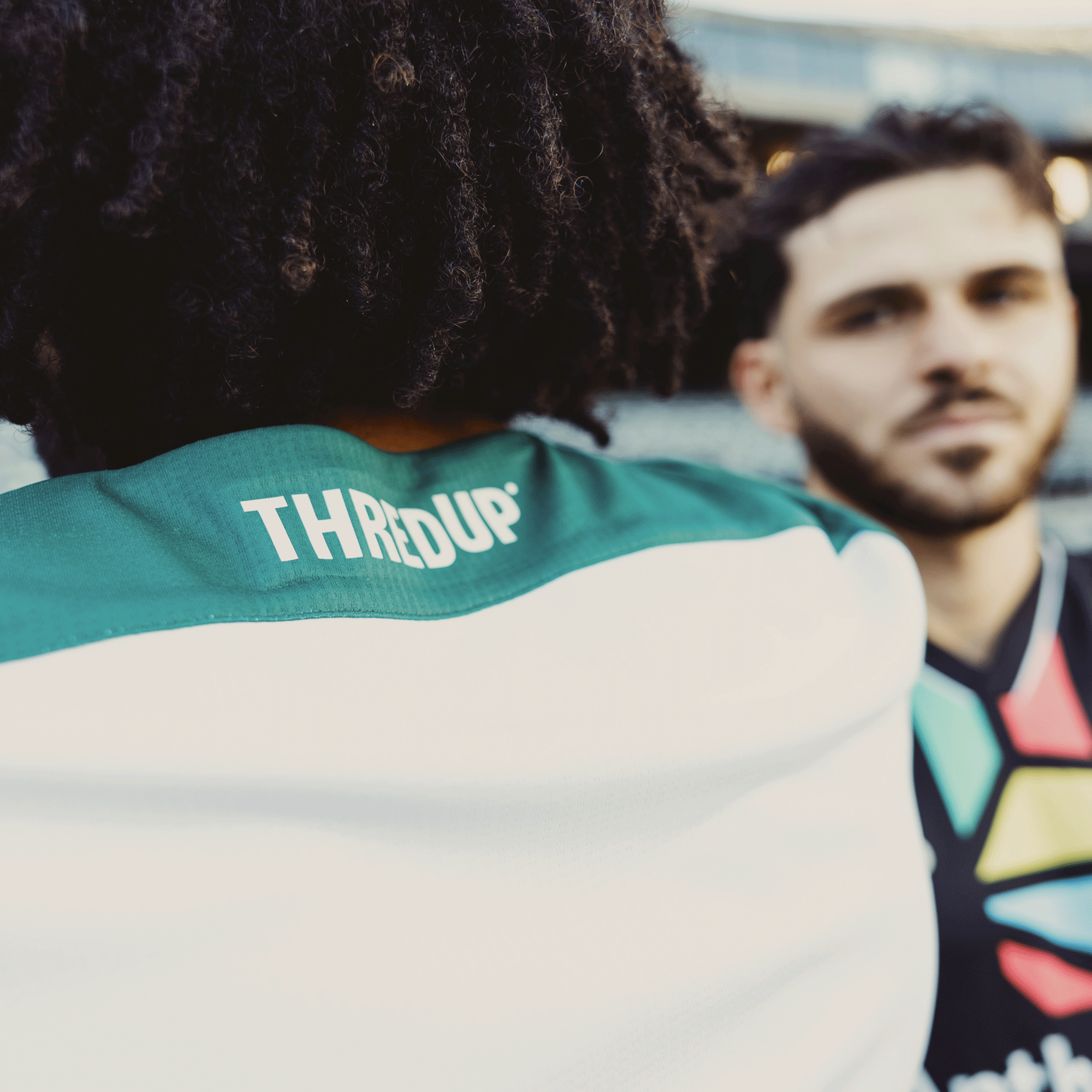 A blurred photo features two people; one with curly hair in an "Oakland Roots SC 2025 Second Kit ft. Anthem Blue Cross" jersey near the Oakland Coliseum. The other figure is also blurry, enhancing the lively feel of what appears to be a sports event tied to Oakland's sports heritage.