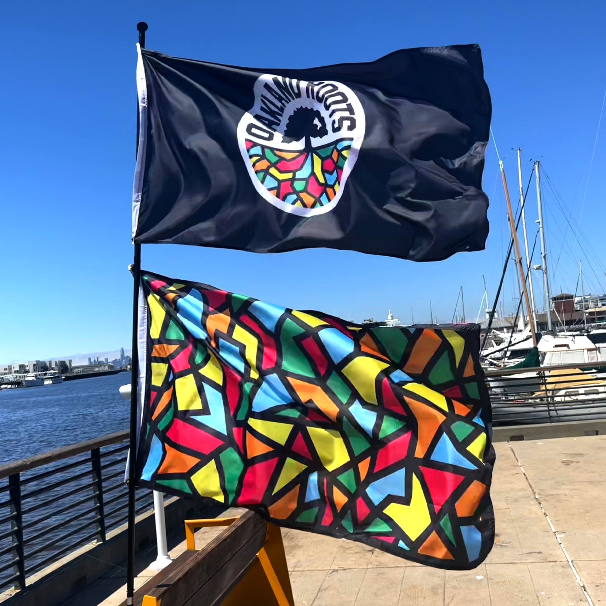 Two colorful flags fly on a pole by a waterfront. The top flag is the Oakland Roots SC Logo Flag, featuring a bold, multicolored Oakland Roots SC logo and the text "Young Roots" on a black background. Below it, another flag showcases a vibrant geometric pattern in red, yellow, green, blue, and black. Boats and a clear blue sky form the backdrop.