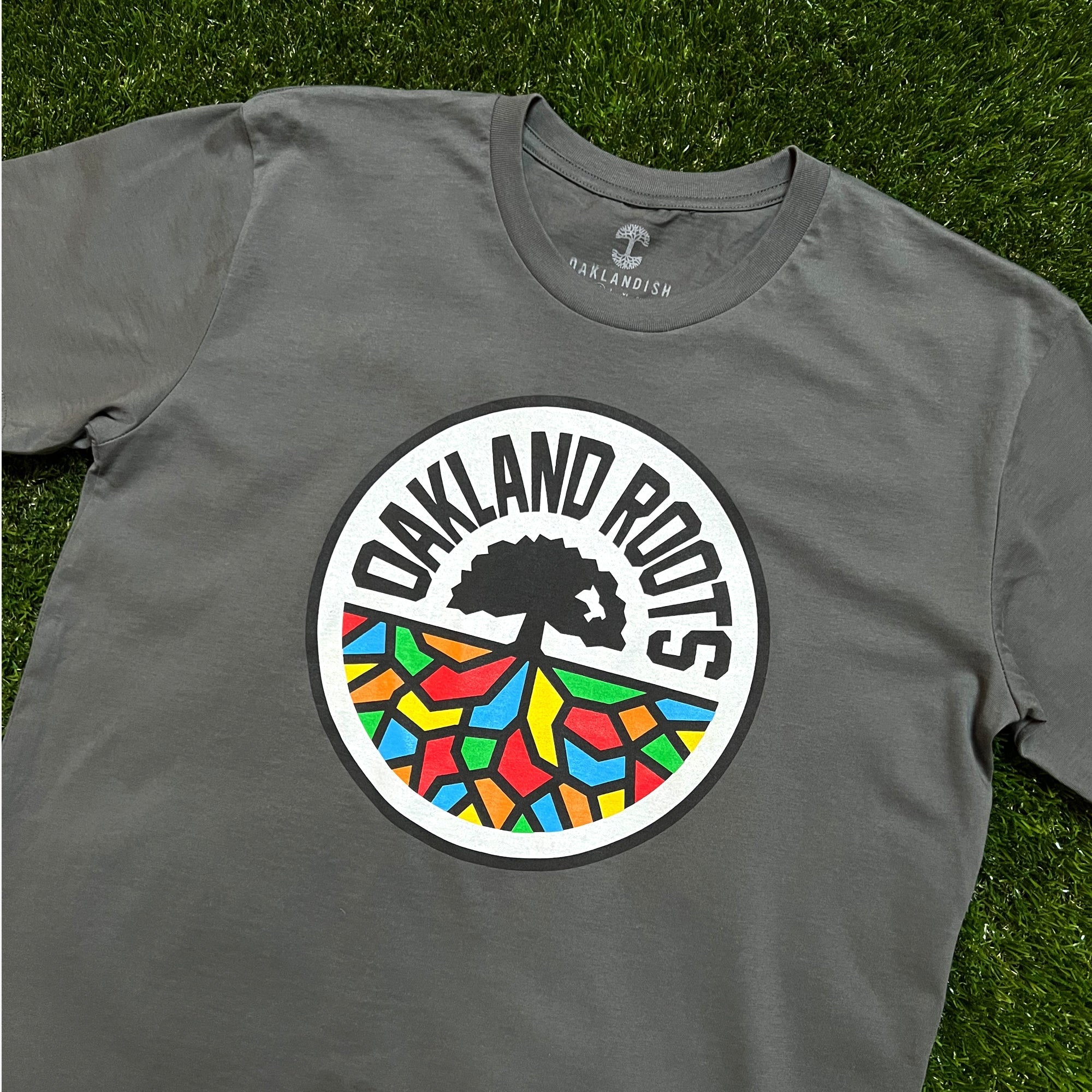 The Oakland Roots SC Classic Tee is a gray t-shirt featuring a striking circular logo at the center. The logo showcases a black tree silhouette above a vibrant pattern of geometric shapes, capturing the essence of the Oaklandish spirit. Bold black letters arching above the tree spell "OAKLAND ROOTS." The tee from Oakland Roots SC is laid flat on green grass.