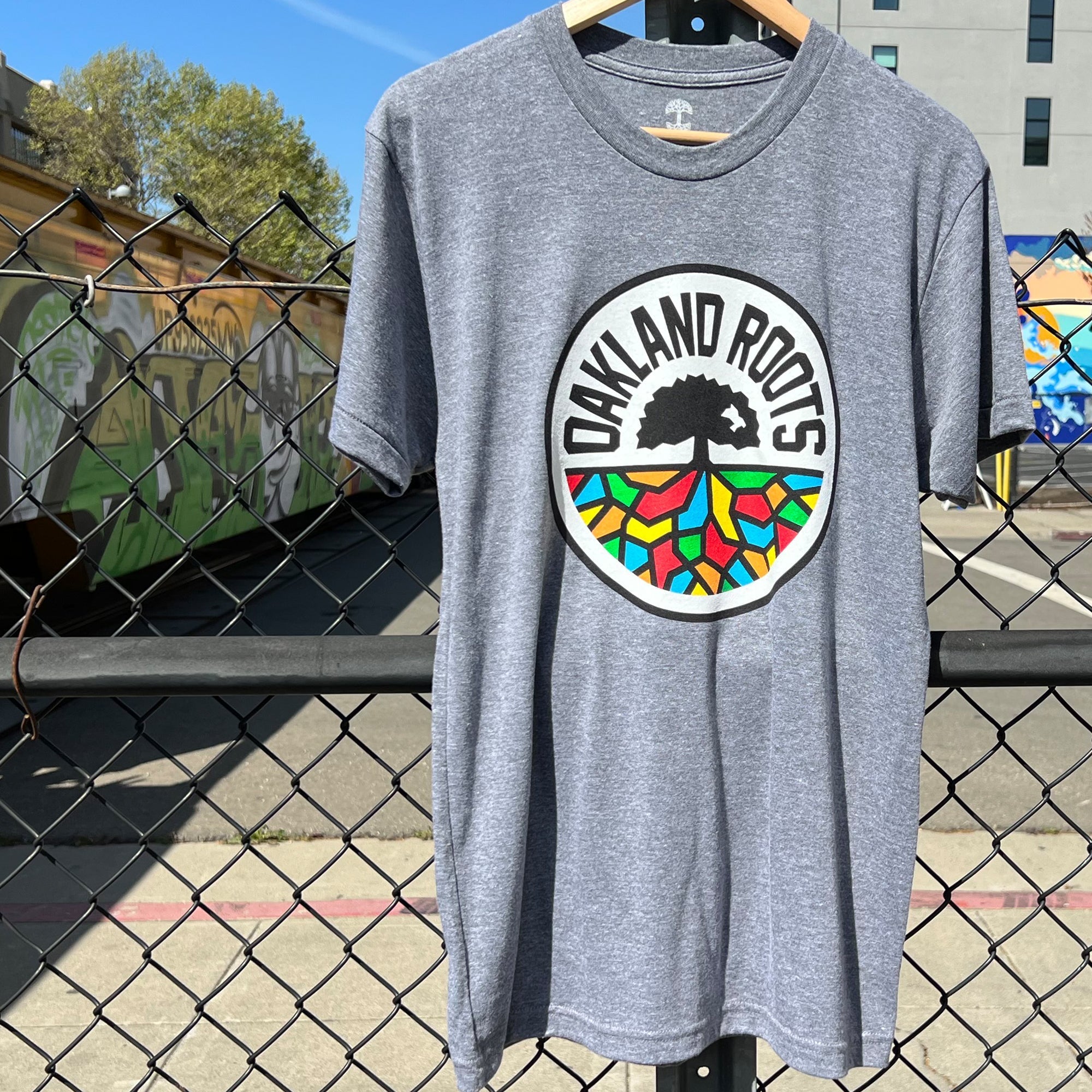 An Oakland Roots SC Classic Tee in gray hangs on a black chain-link fence, highlighting the logo of the Oakland Roots Sports Club, which features "Oakland Roots" encircling a tree graphic with multicolored roots. The backdrop includes a train car adorned with murals, a sidewalk, and part of a building featuring vibrant street art, epitomizing social good.