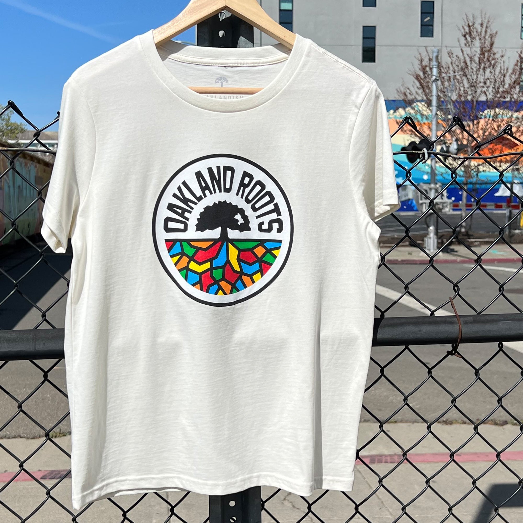 A Women's Oakland Roots SC Classic Tee is displayed on a wooden hanger in front of a chain-link fence, highlighting the "Oakland Roots" logo with a stylized tree and vibrant mosaic base. In the background, an Oakland street reveals a mural and buildings under a clear blue sky—celebrating the spirit of this unique sports club.