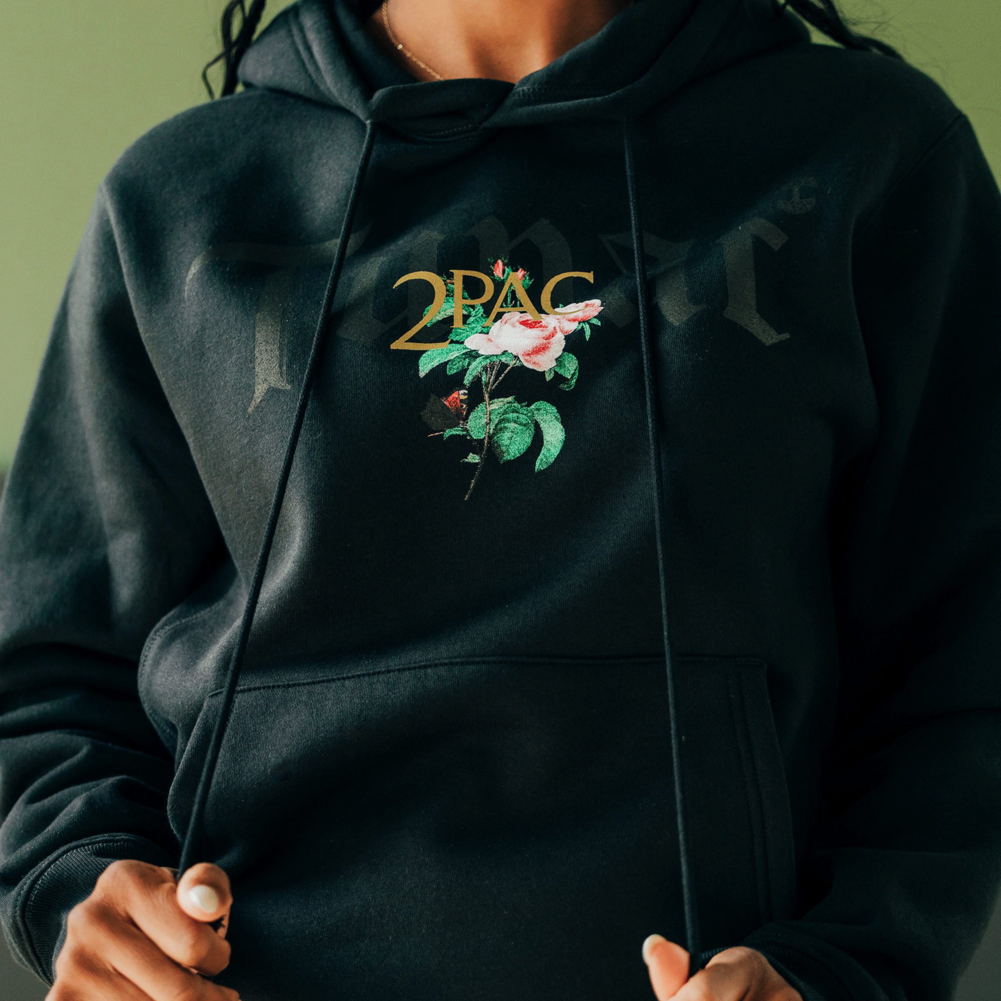 Wearing the Oaklandish Roses Hoodie, which prominently displays a colorful rose and leaves graphic along with "2PAC," a person embodies Tupac's legacy. Against a green background, they adjust the hoodie strings as part of this unique Oaklandish x Tupac hip-hop collection.