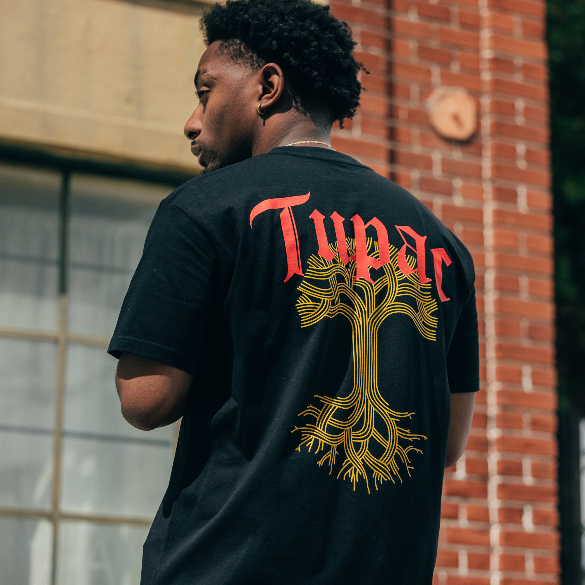 A man is outside in front of a brick building with large windows and a light-colored ledge, wearing the Oaklandish Roses Tee, which is part of the Oaklandish x Tupac collaboration. It features "Tupac" in red lettering above a yellow tree design. He has short curly hair and is turning slightly to the side.