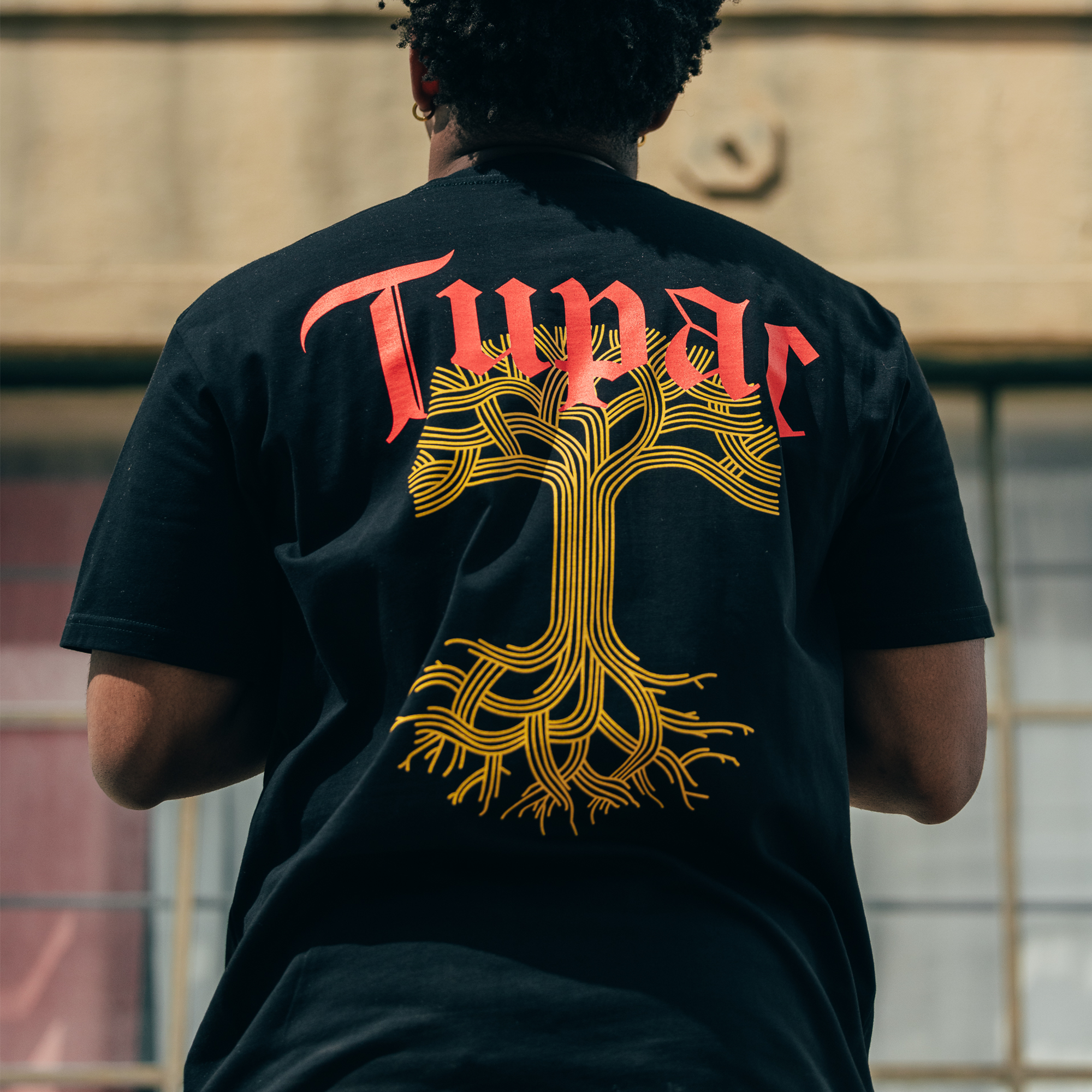 A person is wearing the Roses Tee from Oaklandish, featuring bold red "Tupar" text above a stylized yellow tree graphic, evoking a sense of an Oaklandish x Tupac collaboration. The individual, with curly black hair, stands outside a beige building with large windows, showcasing the T-shirt's design from behind.