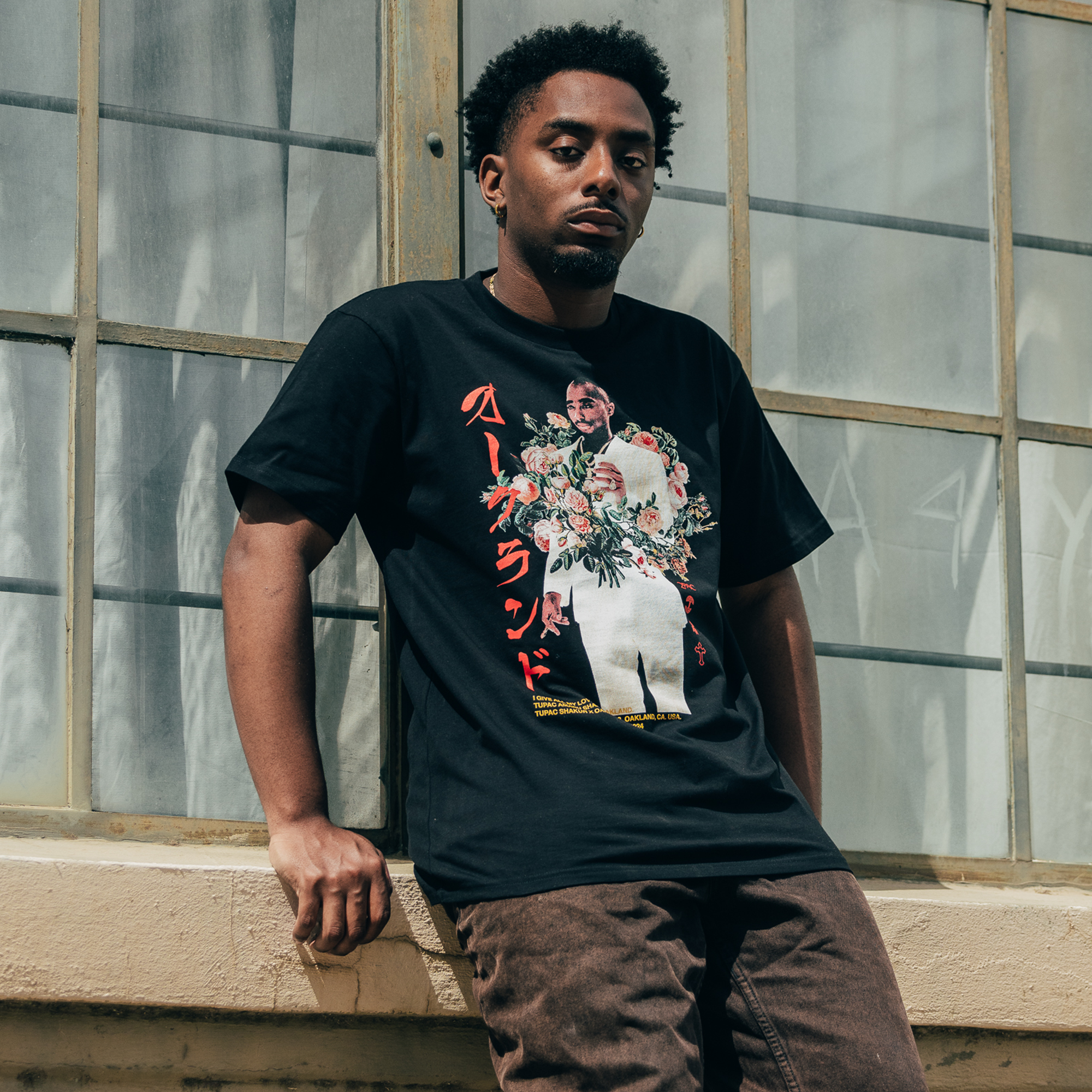 A man leans against a wall in front of large, grid-like windows. Sporting the Oaklandish "Roses Tee," featuring a graphic of a person holding flowers and accented with red Japanese text, he exudes urban flair. With short hair and brown pants, the image captures a casual vibe under soft lighting.