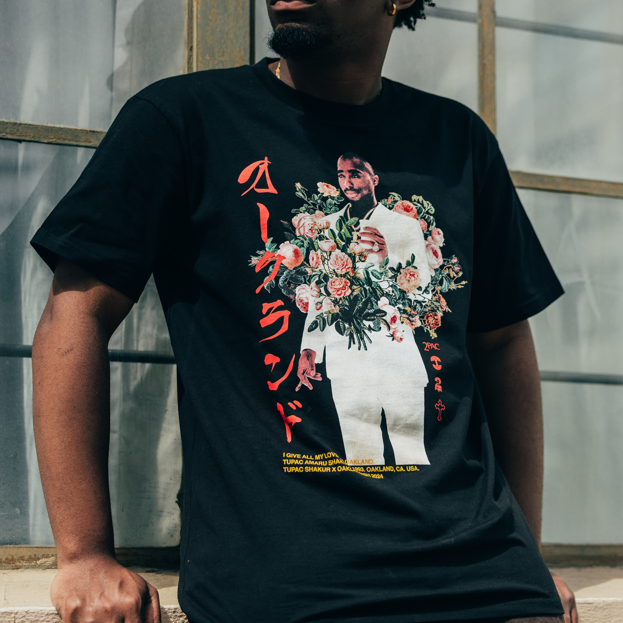 A person wearing the Roses Tee from Oaklandish, featuring a design with a graphic of a man in a white suit holding flowers and red Japanese text. The individual sits in front of a window with crossed arms, facing slightly away from the camera.