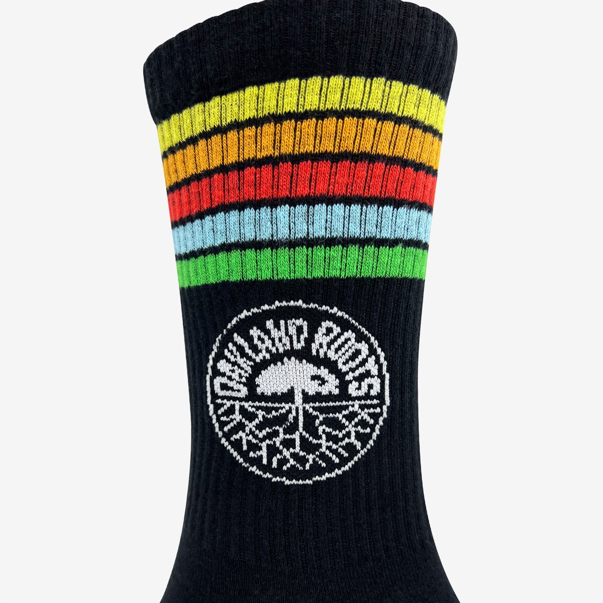 Close-up of an Oakland Roots SC Stripes Sock in black, featuring colorful horizontal stripes at the top in yellow, orange, red, blue, and green. Below the stripes is a white circular logo with a tree and spreading roots prominently displayed. The side text within the circle reads “LOCAL HOOPS.” Perfect for any sports club member.