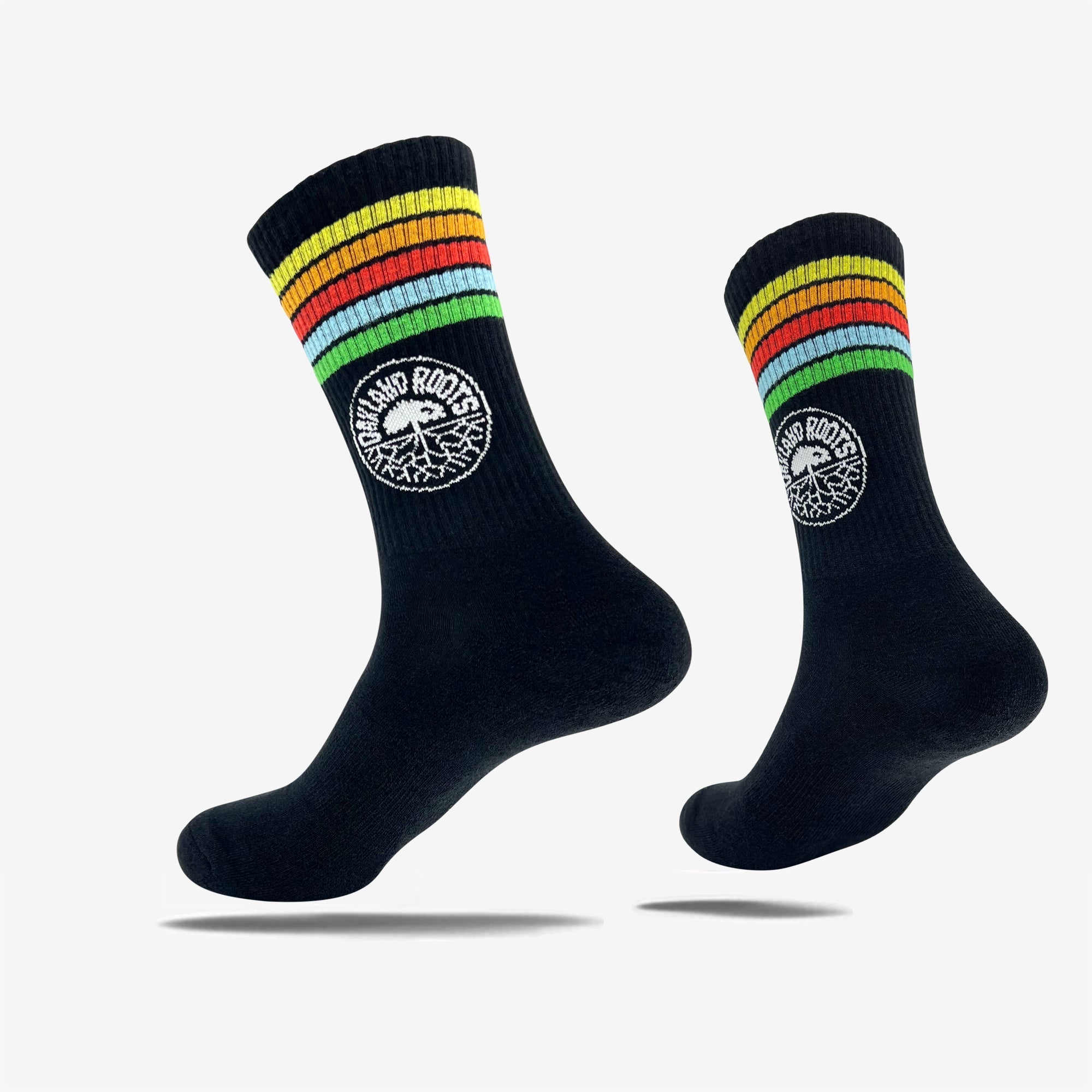 Two black crew socks by Oakland Roots SC, named the Oakland Roots SC Stripes Sock. These feature colorful stripes of red, yellow, green, and blue around the top. Each sock has a white circular logo near the ankle with "Island Roots" text circling a stylized tree design. Perfect for any sports club supporter. The socks are displayed against a white background.