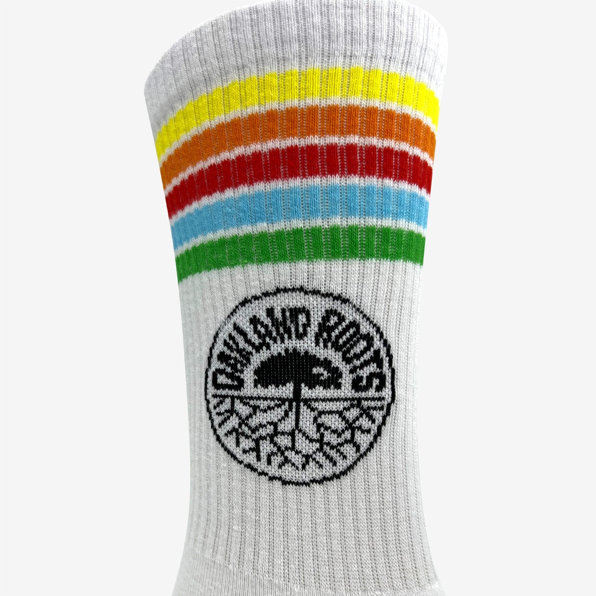 Close-up of the Oakland Roots SC Stripes Sock, a white crew sock from Oakland Roots SC. It showcases horizontal rainbow stripes in vibrant shades of yellow, orange, red, blue, and green near the top. Below the stripes sits a black circular emblem that displays the text "Oakland Roots" encircling an illustration of a palm tree with visible roots.
