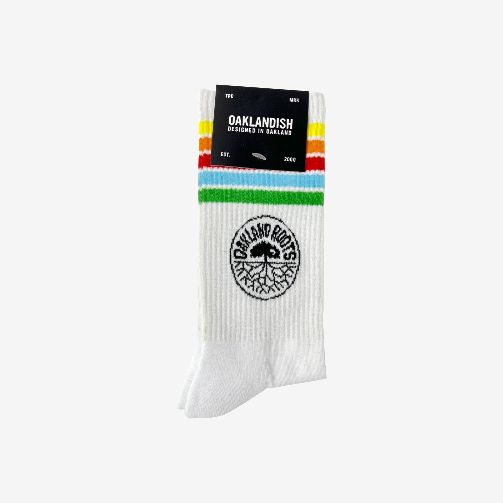 Introducing the Oakland Roots SC Stripes Sock: white athletic crew socks featuring a black tree emblem and "OAKLAND ROOTS" text. These stylish socks are adorned with green, yellow, and red stripes near the top. They come with a sleek black cardboard label displaying "OAKLANDISH," "Designed in Oakland," and "Est. 2000" in white text. Available in size MED.