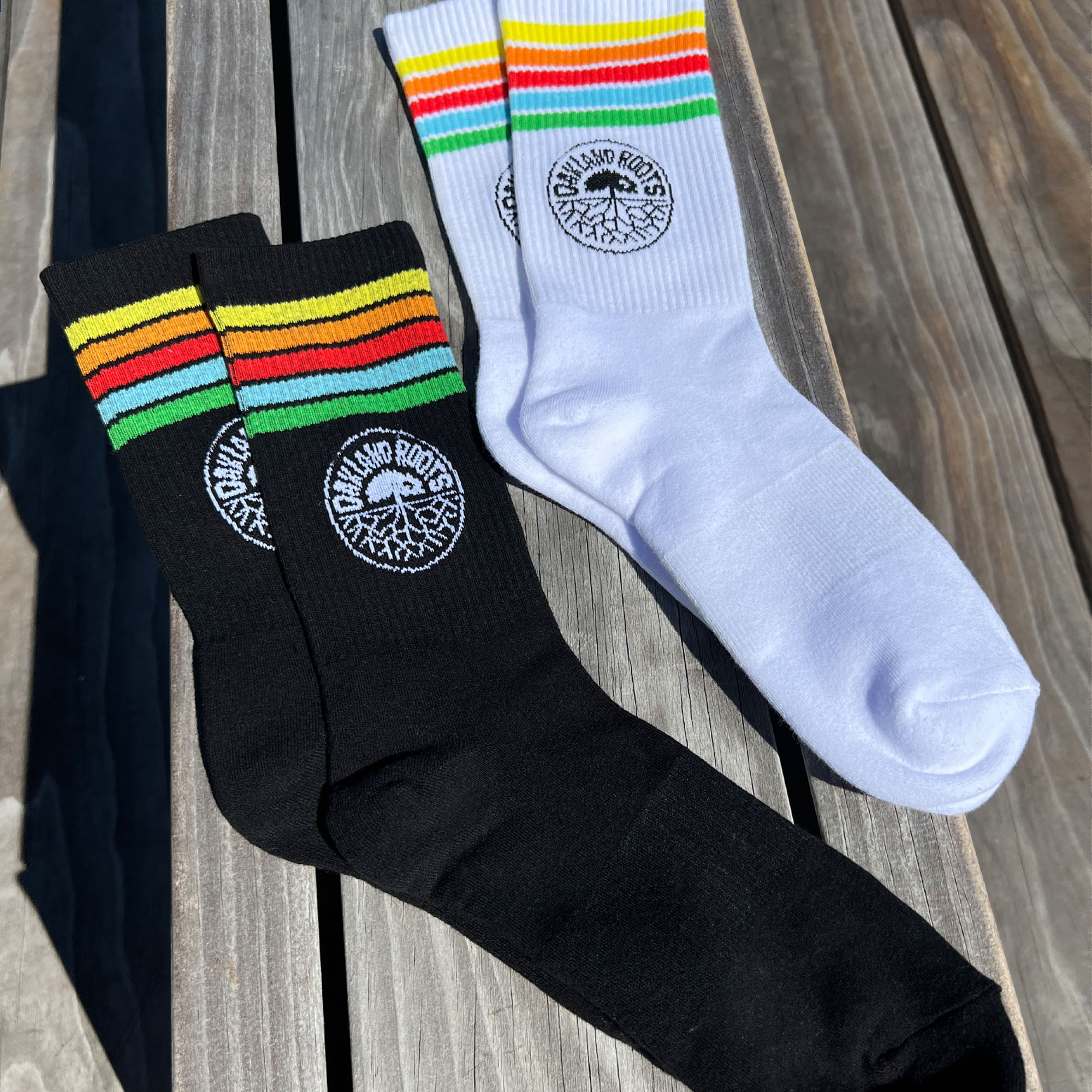 Two pairs of Oakland Roots SC Stripes Socks from Oakland Roots SC are displayed on a wooden surface. One pair is black with red, yellow, and green stripes at the top, while the other is white with the same stripe pattern. Both pairs feature a circular Oakland Roots logo with text and a stylized tree at the ankle area. The socks are neatly arranged side by side.