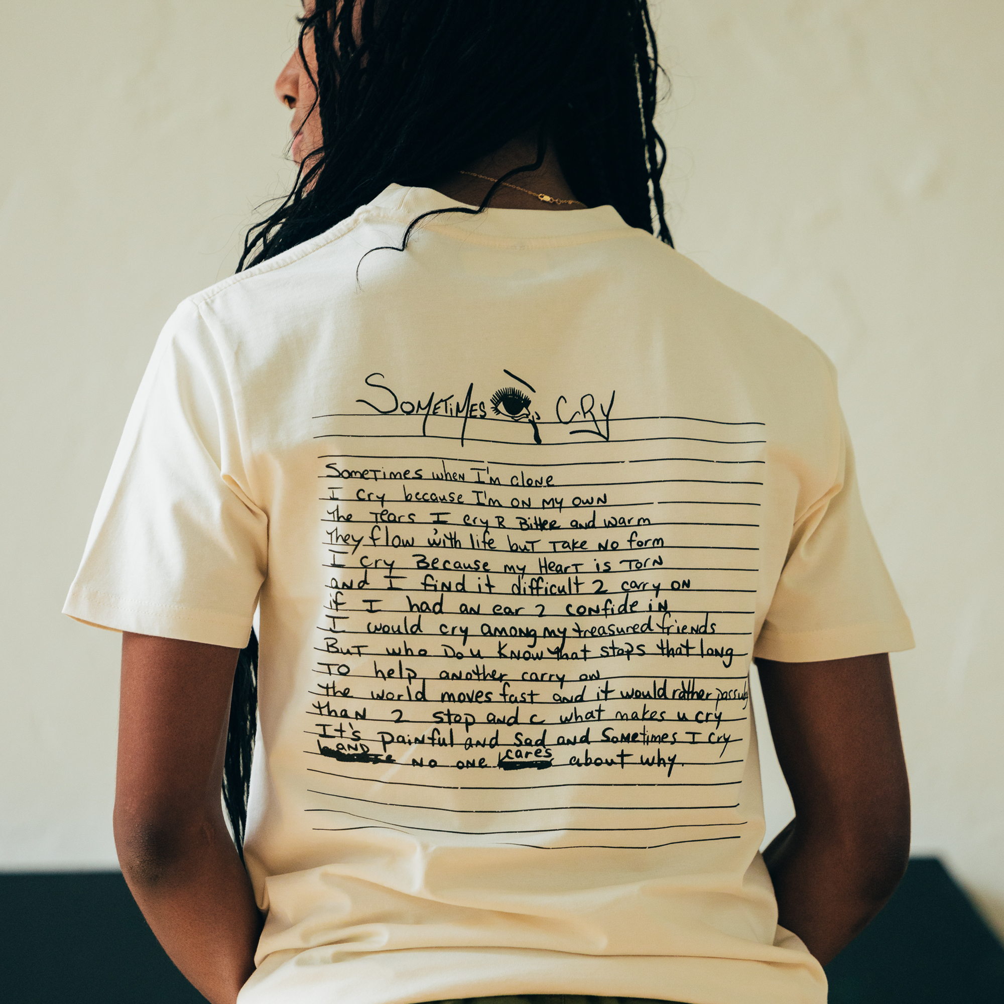 A person with long braided hair stands outdoors against a blurred backdrop, wearing the Oaklandish "Sometimes Eye Cry Tee." This white T-shirt features handwritten text and an illustration of an eye shedding a tear, encapsulating the emotional depth inspired by Tupac Shakur's lyrics. The scene evokes an urban hip-hop vibe as they face away from the camera.