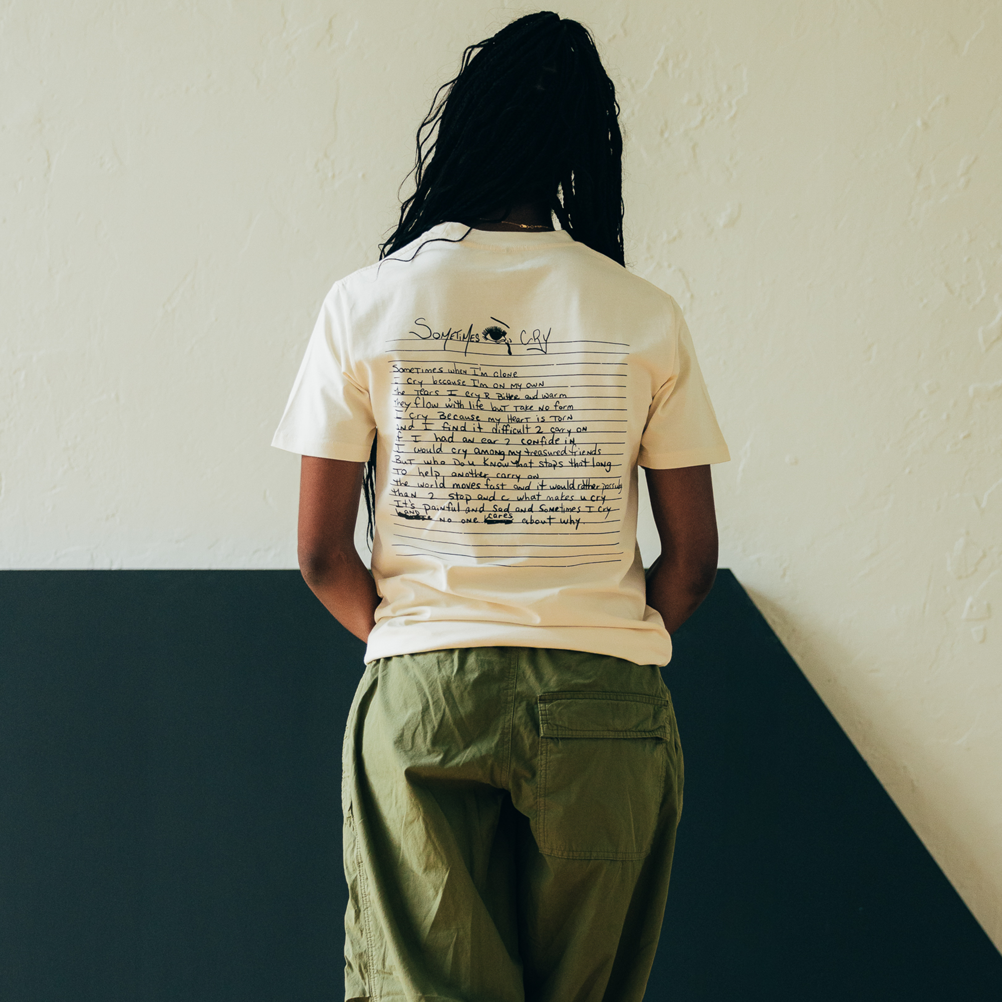 A person with long braids is facing away, dressed in the "Sometimes Eye Cry Tee" by Oaklandish—a cream-colored T-shirt featuring handwritten text and a small graphic that channels Oaklandish street style on the back. They are also wearing olive green pants. The backdrop is a light-colored wall with a dark geometric design, reflecting hip-hop vibes.