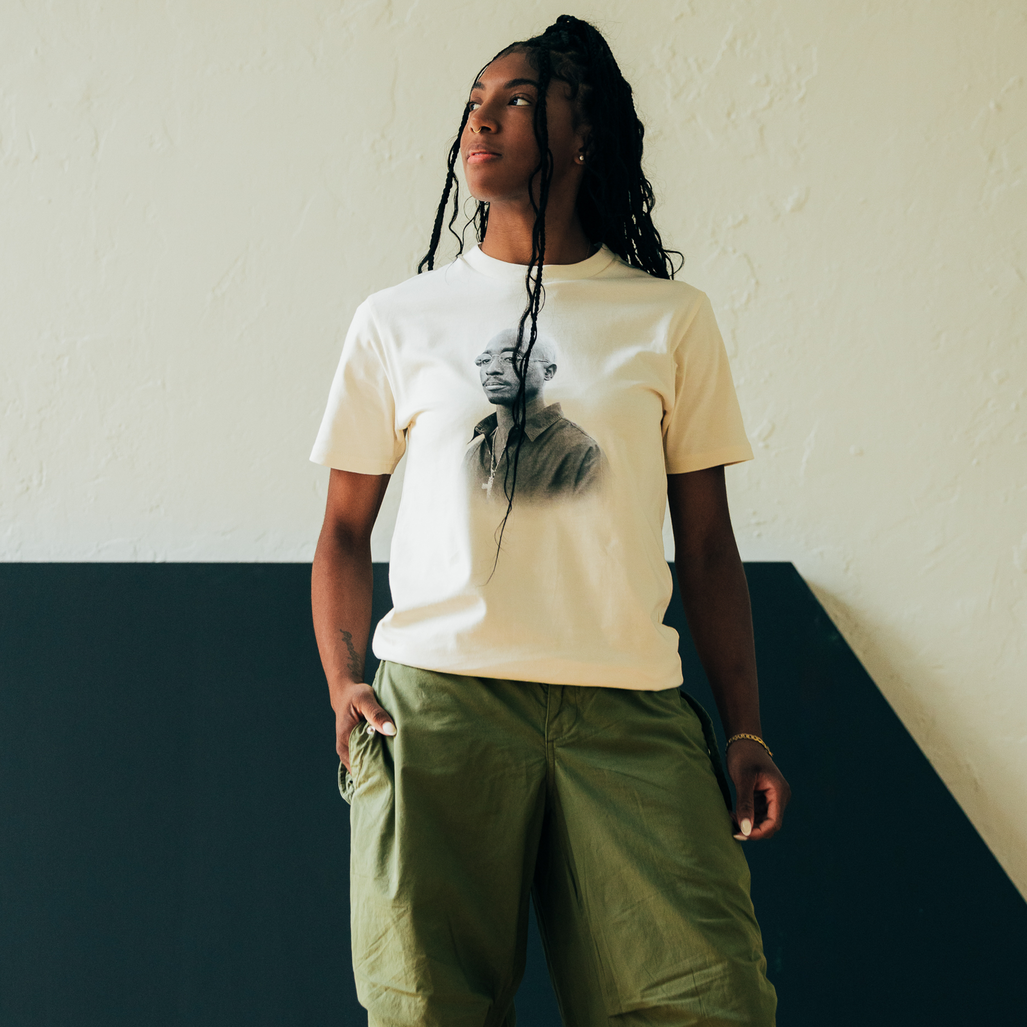 A person with long braided hair stands confidently against a two-tone wall, exuding hip-hop vibes. They wear an Oaklandish Sometimes Eye Cry Tee featuring a black-and-white portrait of Tupac Shakur and loose-fitting green pants. One hand in their pocket, their head is turned slightly to the side.