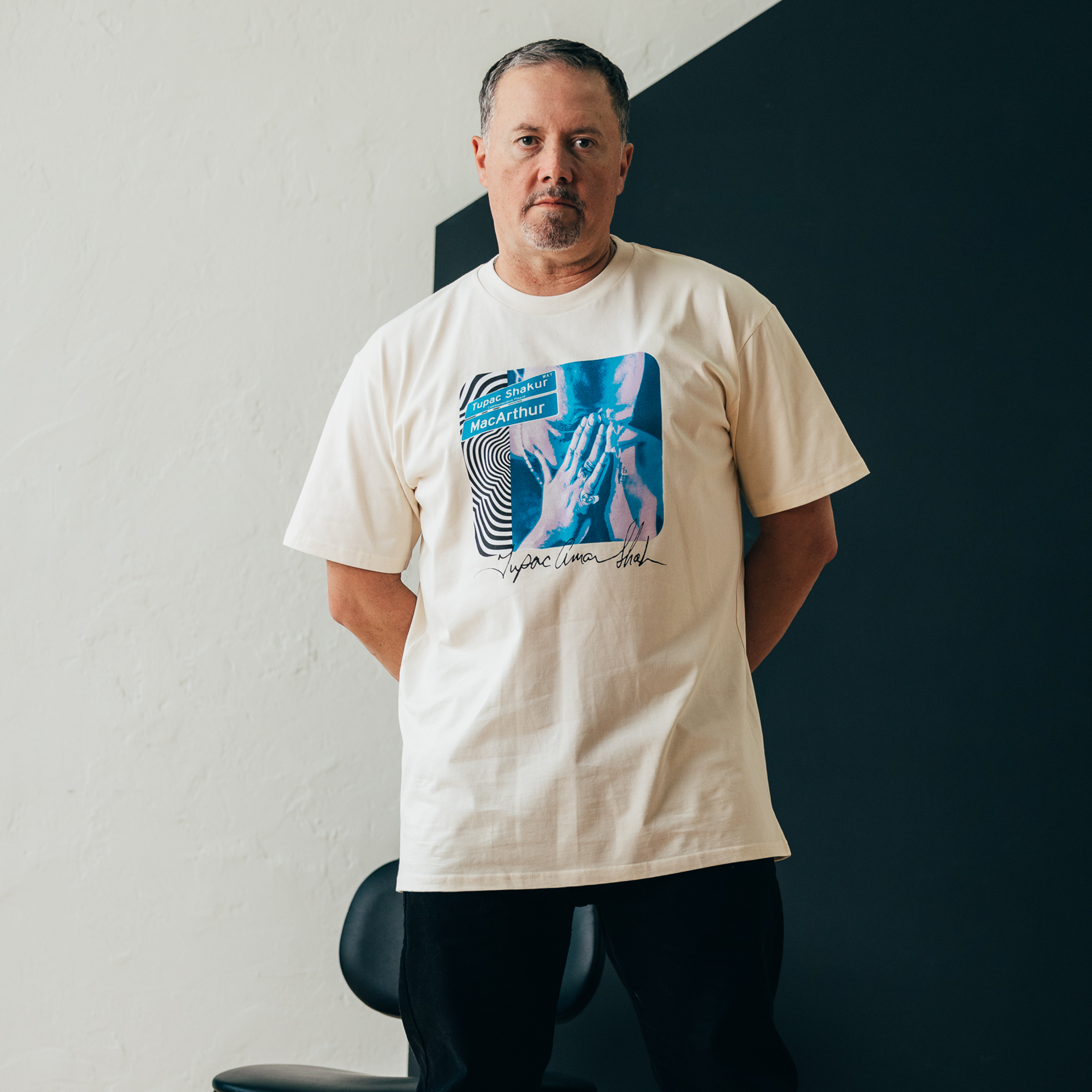 A man stands in front of a black and white wall, wearing the Wavy Tupac Tee from Oaklandish, which features a cream color with a colorful graphic design of abstract shapes and text. Channeling his inner Tupac, he radiates a cool hip-hop vibe. With short gray hair and a neutral expression, his hands are casually clasped behind his back.