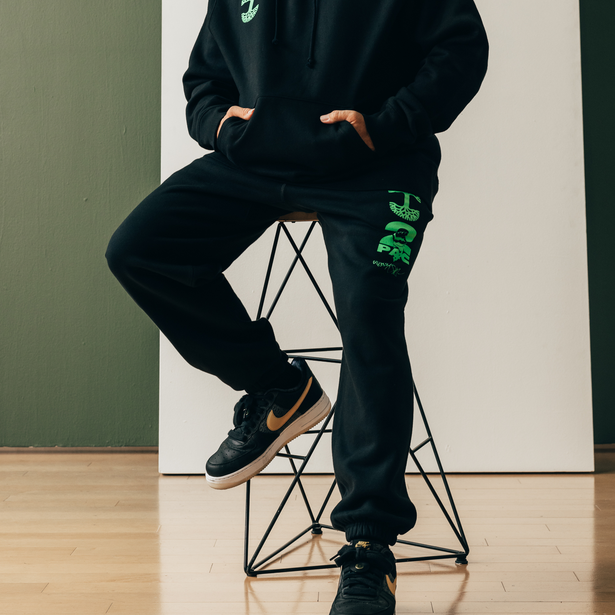 A person sits casually on a black stool wearing a black hoodie and the Oaklandish You Got To KYHU Jogger, both showcasing neon green graphics. The outfit is complemented by black sneakers with a yellow Nike logo. A green wall and white panel backdrop, along with the wood floor, create contrast in the scene.