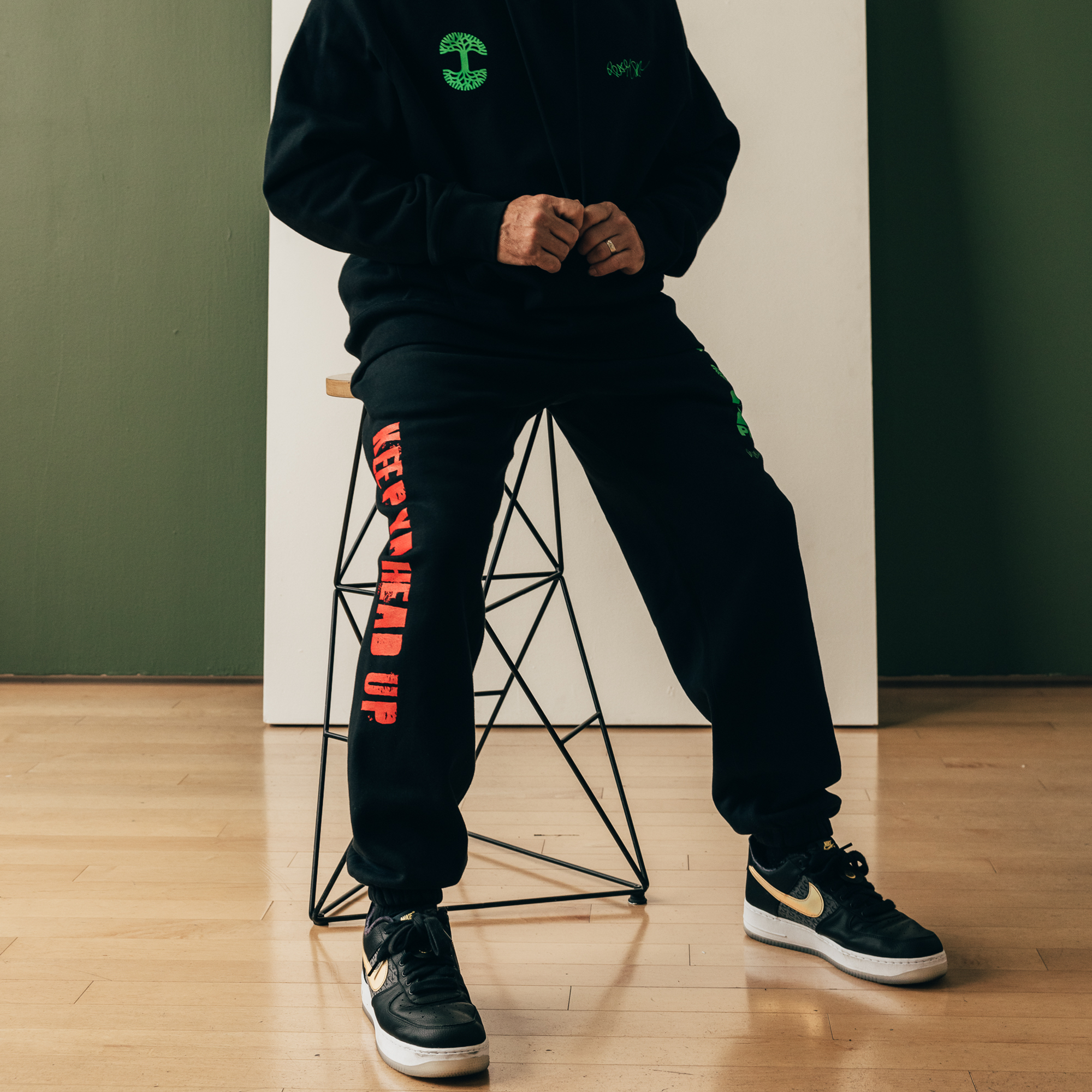 A person sits on a geometric metal stool, sporting a black hoodie and the You Got To KYHU Jogger from Oaklandish's hip-hop collection. The joggers feature red text stating "KEEP YOUR HEAD UP," paying homage to the Tupac collaboration. They complement their look with black and white sneakers adorned with the swoosh logo, set against a backdrop of a white panel and green wall on wooden flooring.