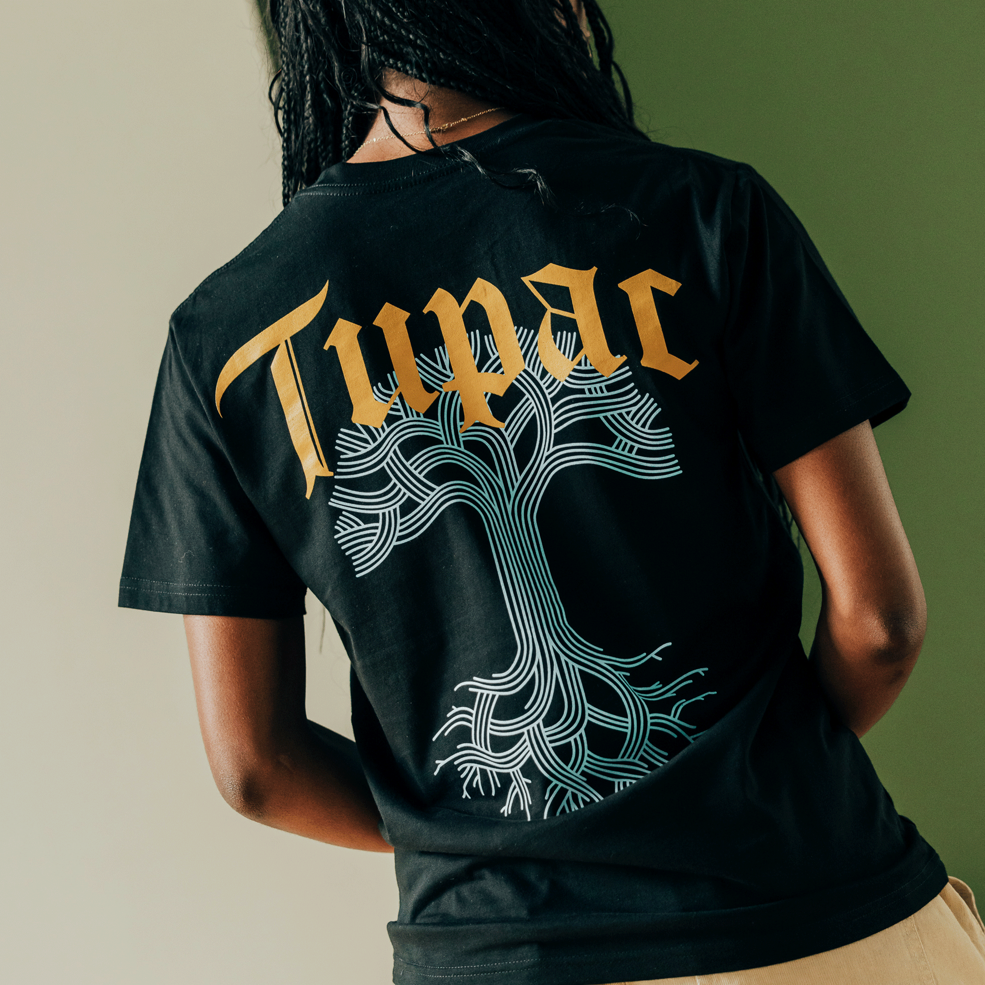 A person with braided hair is wearing the iconic "Throne Tee" by Oaklandish, featuring "Tupac" in bold gold letters above a tree graphic that transforms its intricate white branches into the letter "T," capturing Oakland's hip-hop vibe against a half green and half light gray backdrop.