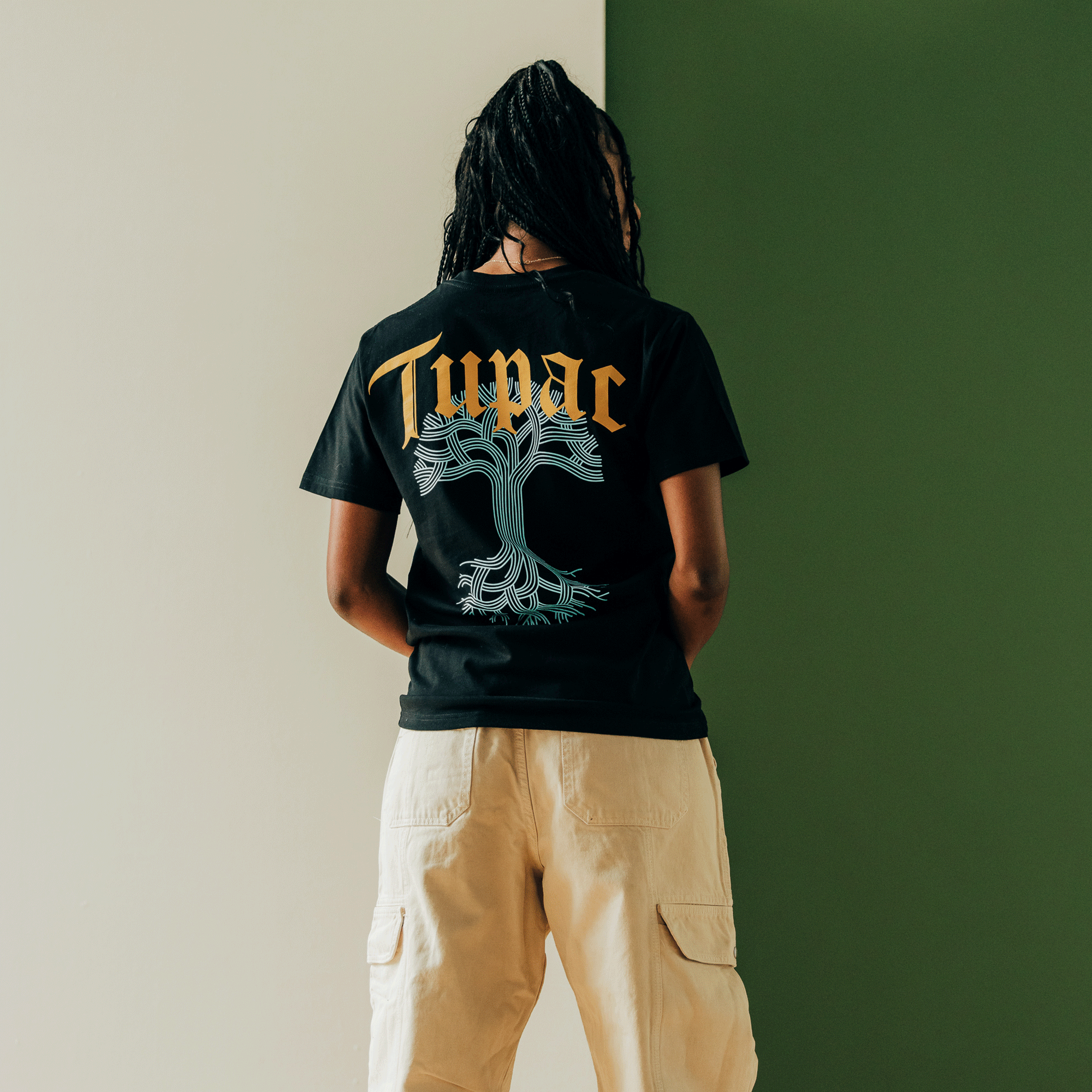 The individual, sporting lengthy dark braids and posing against a backdrop divided into white and green sections, is wearing an Oaklandish "Throne Tee." This black t-shirt features gold "Tupac" text alongside a blue tree graphic and is paired perfectly with beige cargo pants to capture the hip-hop vibe.