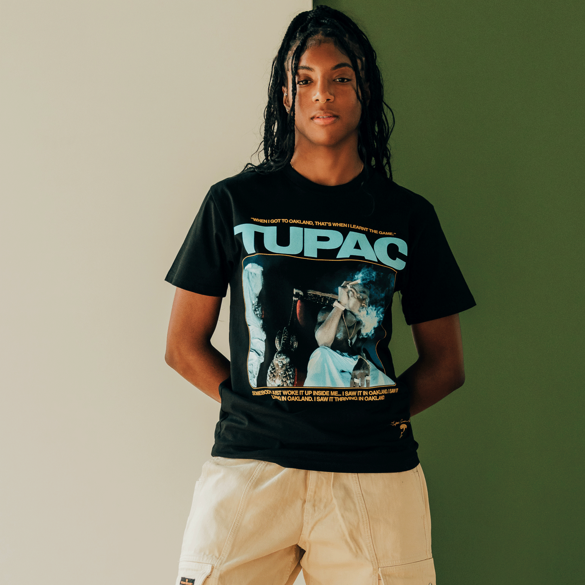 In front of a green wall, someone is wearing the Throne Tee from Oaklandish. This black T-shirt features "Tupac" in large blue letters, celebrating the legendary hip-hop icon. It's decorated with images and text, making a perfect match with light-colored shorts. The individual has long dark hair that echoes Oakland's vibrant style.