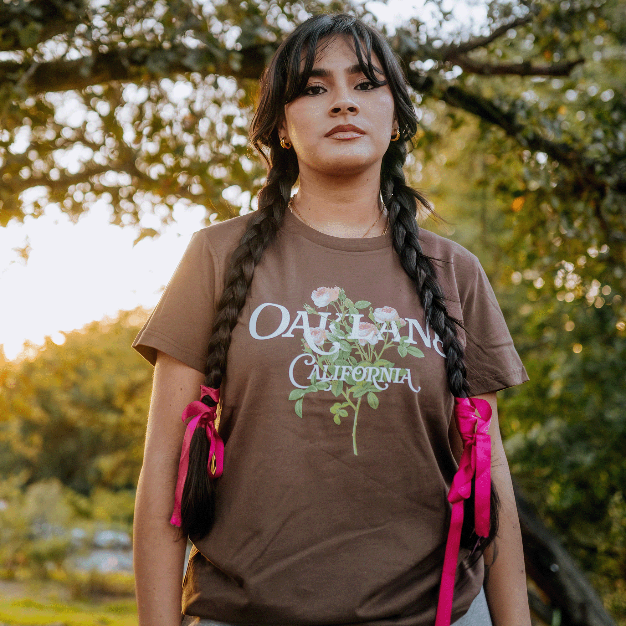 A woman stands outdoors wearing the Women's Posy Tee from Oaklandish, a classic fit 100% cotton T-shirt adorned with "Oakland California" and floral designs. Her long dark hair is styled in braids with pink ribbons. The backdrop of trees and a setting sun casts a warm glow as she gazes into the camera with a calm expression.