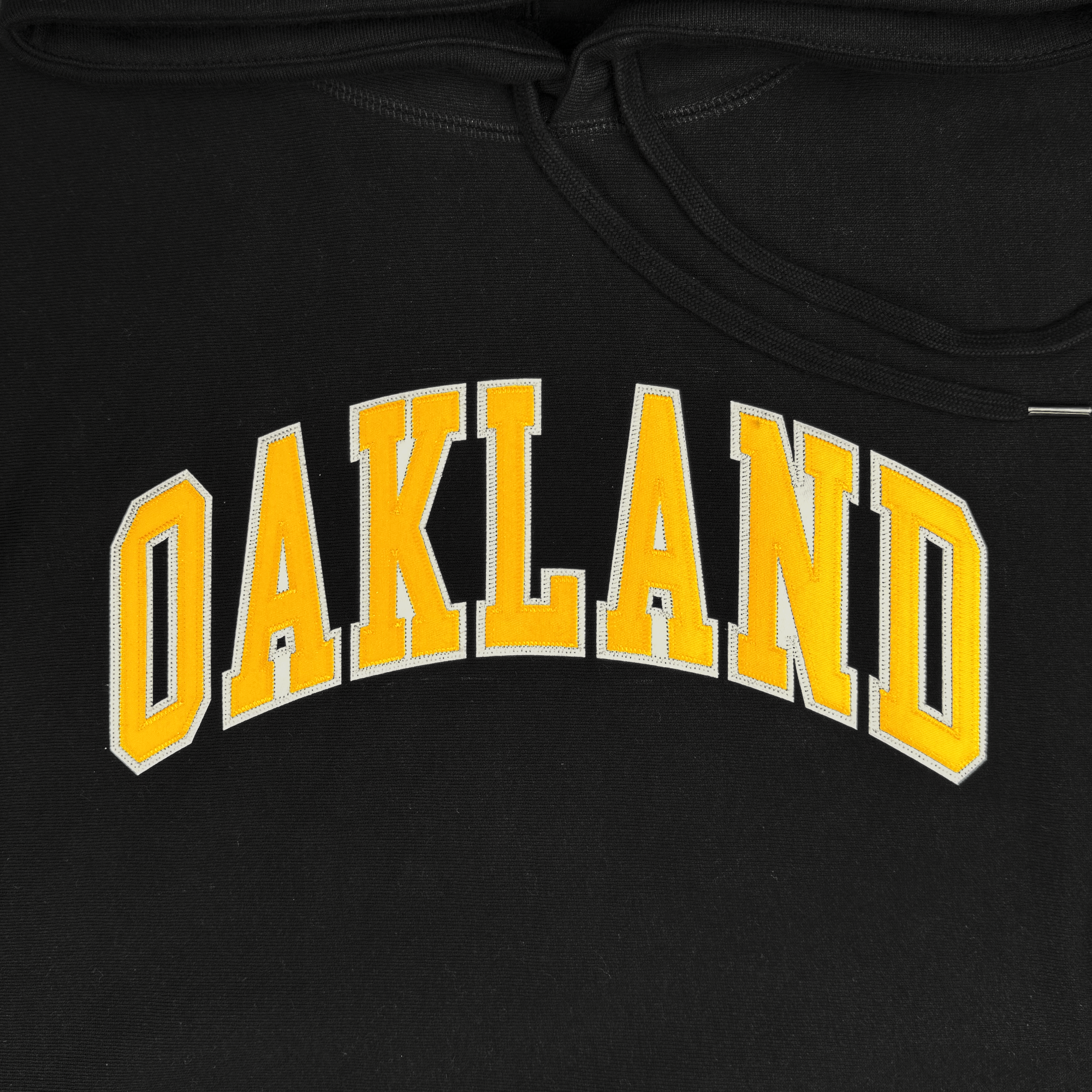 The Alma Mater Heavy Hoodie by Oaklandish is a black hoodie featuring "OAKLAND" in bold yellow letters outlined in white, arched across the chest. Made from thick, durable fabric, it showcases a visible neckline and part of the hood at the top, combining stylish design with cozy comfort.