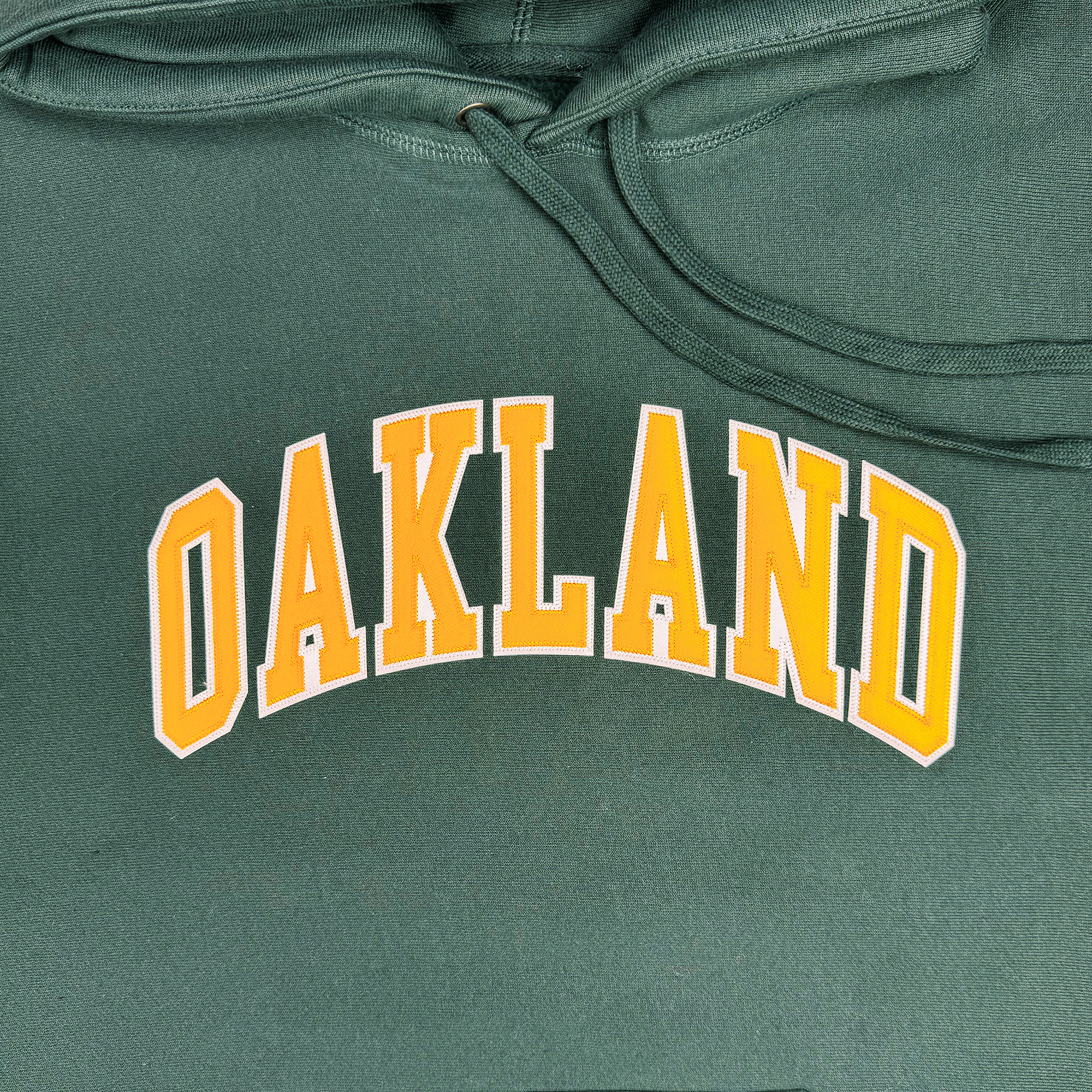 The Alma Mater Heavy Hoodie by Oaklandish is a green fleece hoodie showcasing "OAKLAND" in prominent, bold yellow letters with a white outline, perfectly centered on the fabric. Visible hood strings accentuate the neckline, and the material appears soft and comfortable.