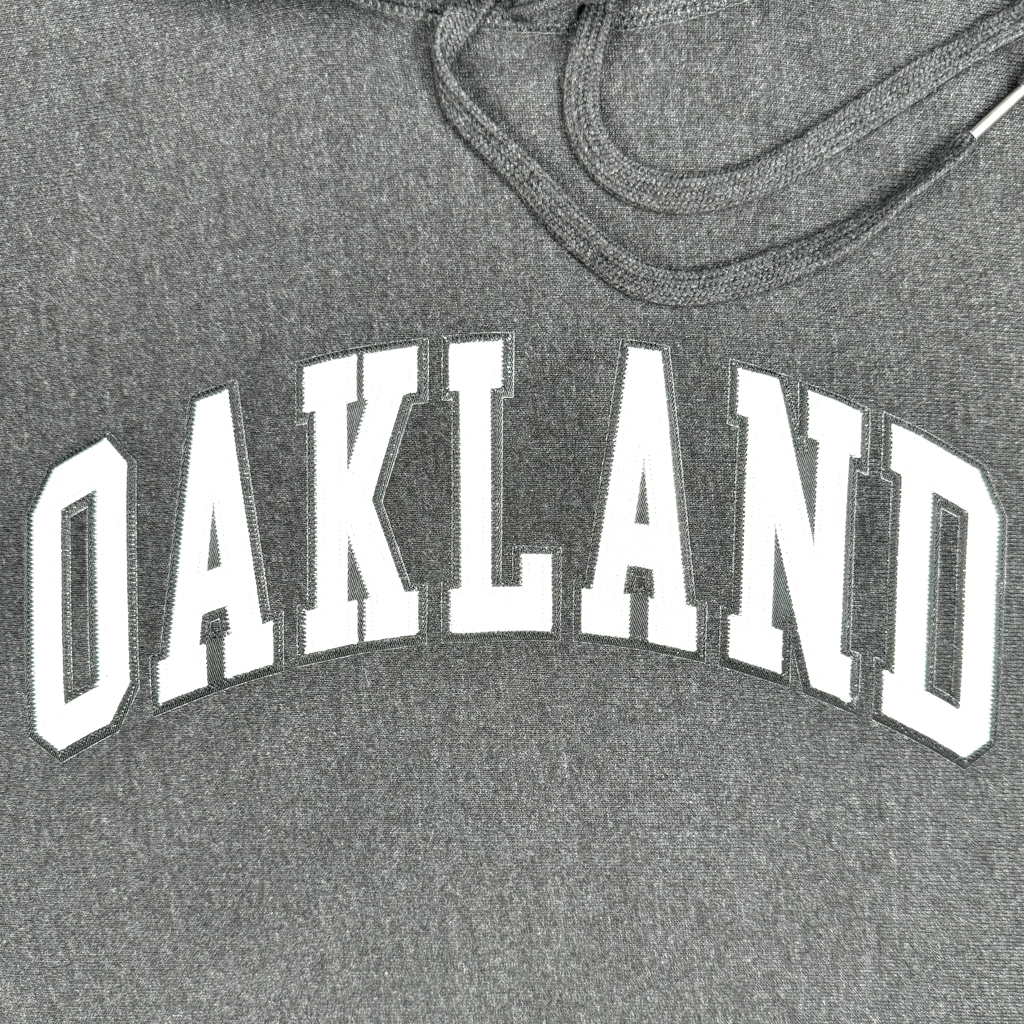 The Alma Mater Heavy Hoodie by Oaklandish is a gray hoodie showcasing "OAKLAND" in bold, slightly arched white capital letters across the chest. It features a drawstring visible from the hood.