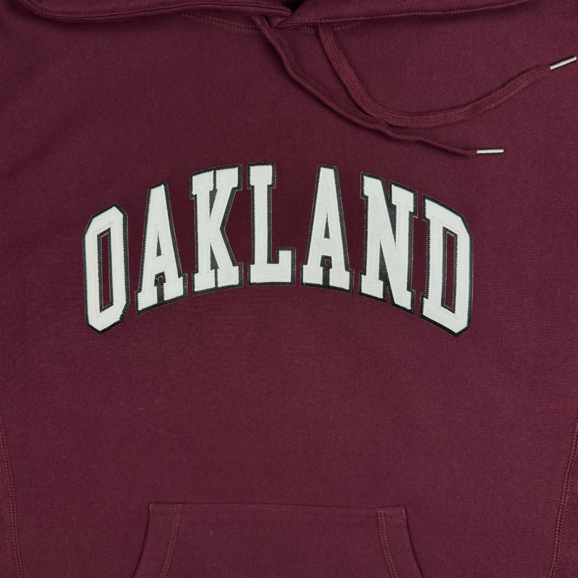 Introducing the Alma Mater Heavy Hoodie by Oaklandish: a maroon pullover hoodie that features "OAKLAND" in bold, white block letters arched across the chest. Made from premium heavyweight fleece, it offers a soft texture with a visible drawstring on the hood and a convenient kangaroo pocket at the bottom.