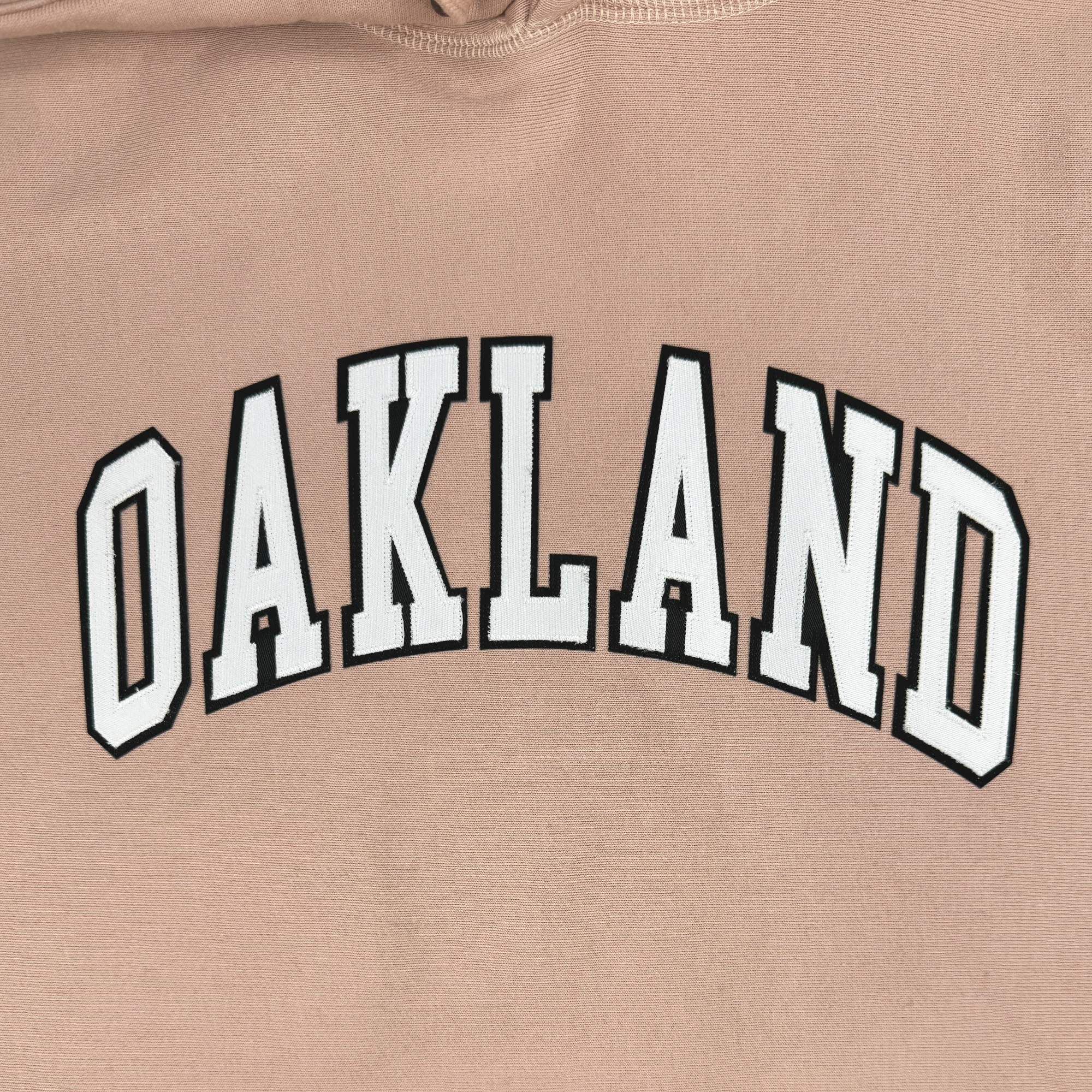 The Alma Mater Heavy Hoodie from Oaklandish is a light brown fleece hoodie that showcases the word "OAKLAND" in bold, uppercase white letters with a black outline. The lettering is elegantly arched and centered across the fabric, merging comfort with style seamlessly.