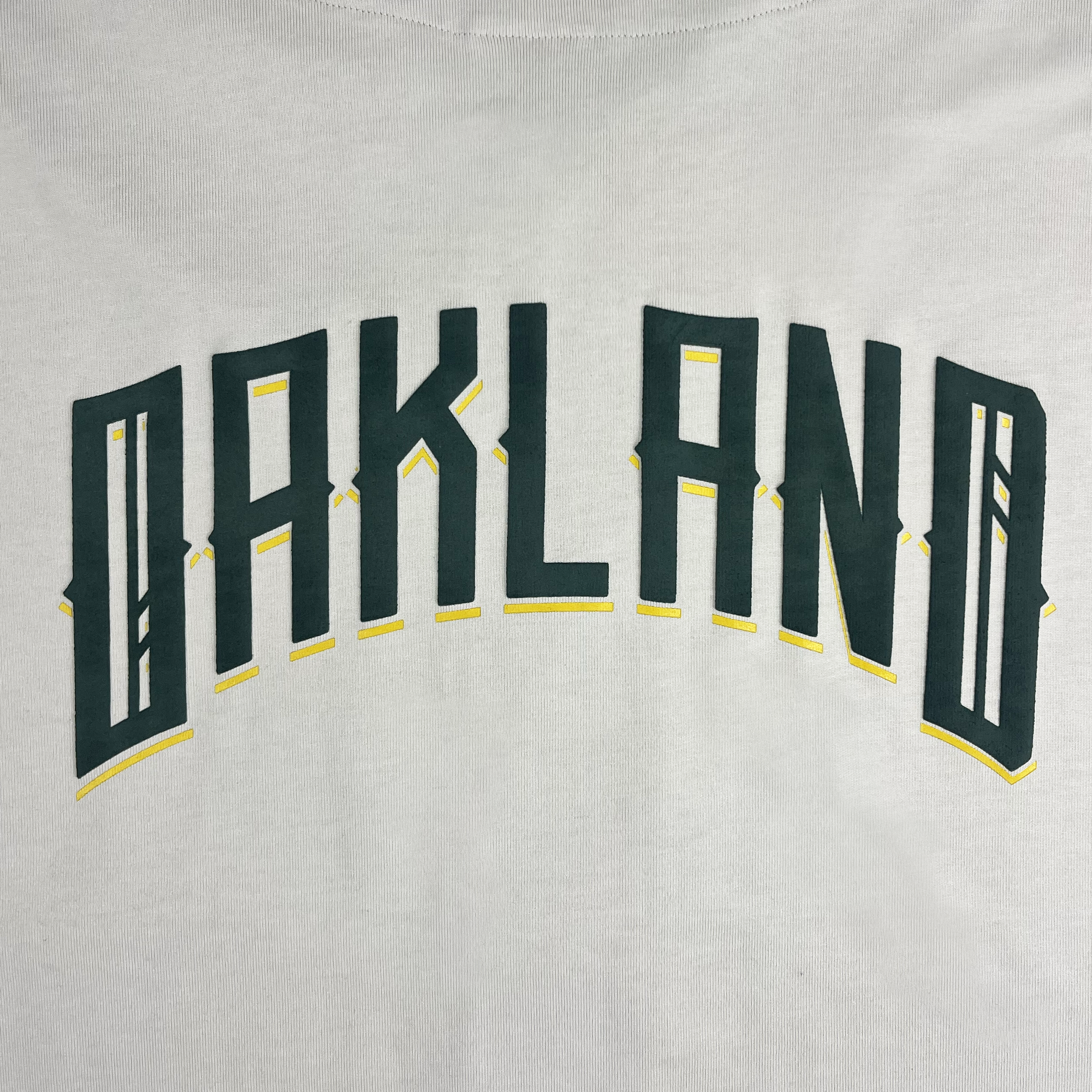 The Oakland Ballers Heavy Baseball Jersey features a white fabric with the word "OAKLAND" emblazoned in bold, dark green capital letters accented with yellow. The text has a stylized and arched design, evoking the sporty aesthetic of the Oakland Ballers, a professional baseball team in the Pioneer League.