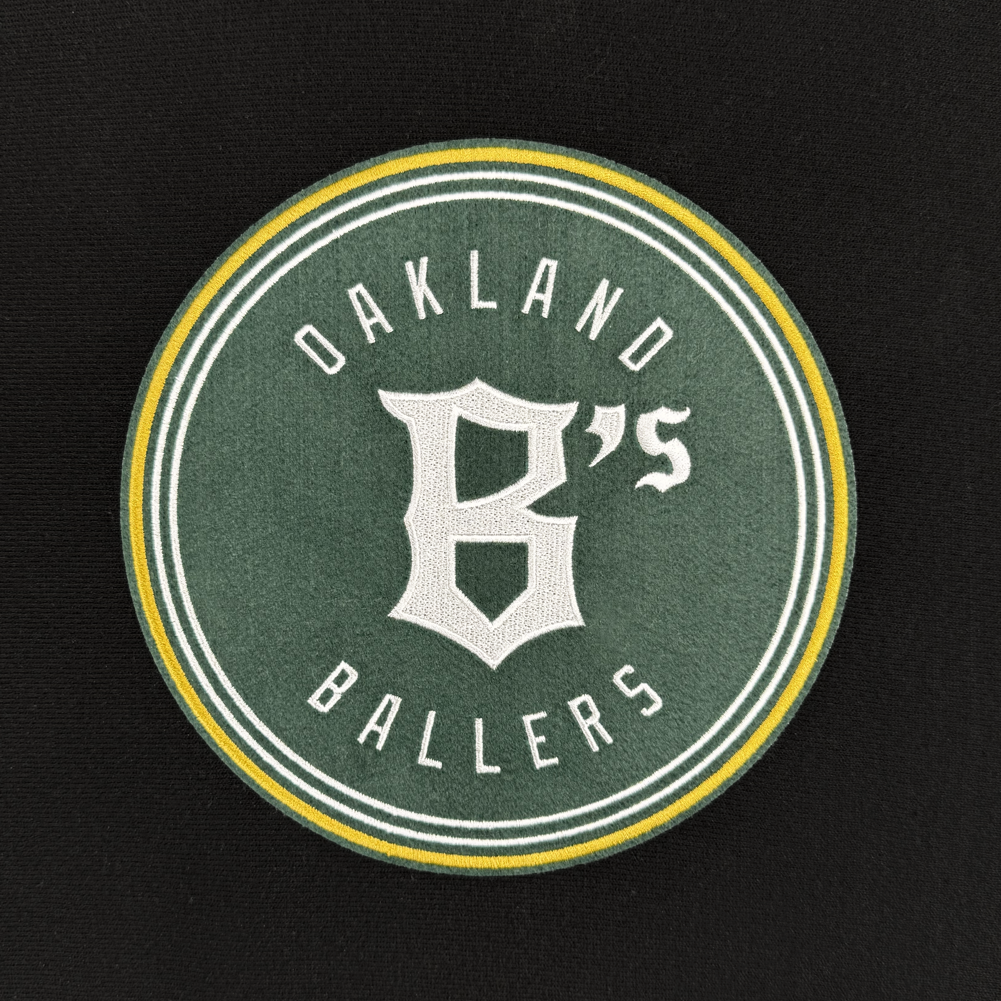 On a black backdrop, a circular patch features the words "Oakland Ballers" in white text encircling a bold stylized "B's" at the center. This green patch, accented with yellow and white borders, honors the Oakland Ballers—a professional baseball team in the Pioneer League—available on the Champion X Oakland Ballers Logo Hoodie.